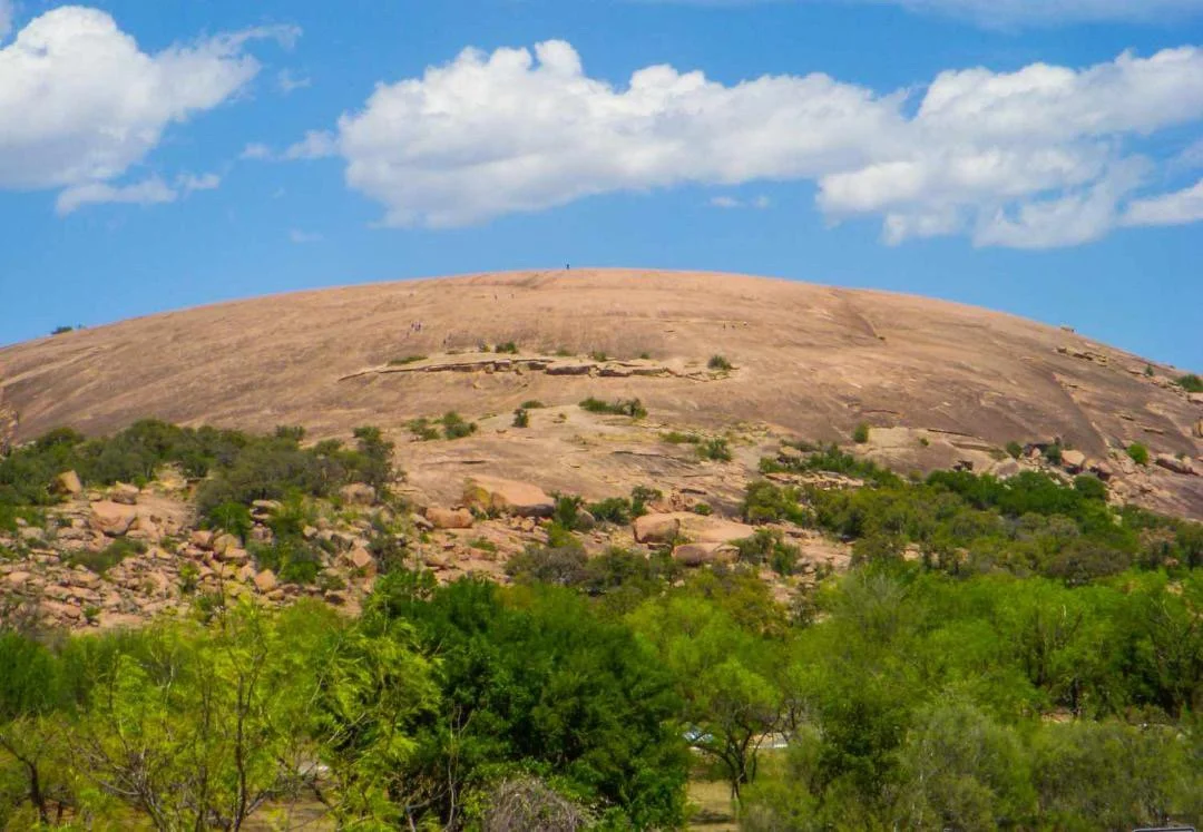 Large, rounded granite hill with sparse vegetation on top, surrounded by green trees under a partly cloudy sky.
