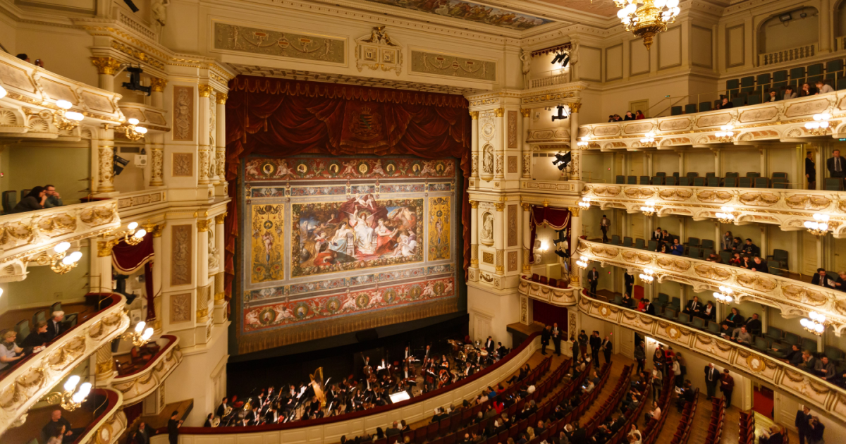 Inside a grand theater with ornate balconies filled with audience, a large decorated stage with a tapestry backdrop, and an orchestra pit with musicians preparing for a performance.