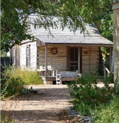 A rustic wooden cabin with a porch, surrounded by greenery and trees, with an outdoor sofa and table on the porch.