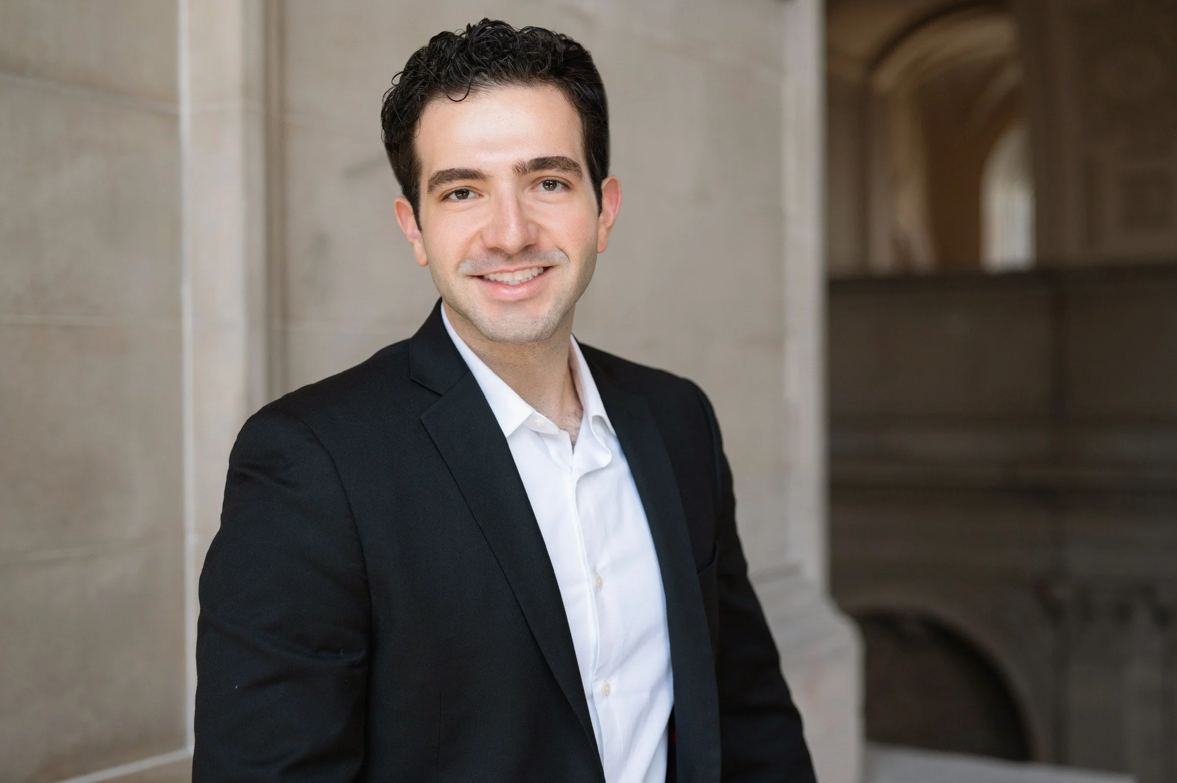 Young man with short curly dark hair, wearing a black suit jacket and white dress shirt, smiling, standing in front of a stone building with arches.