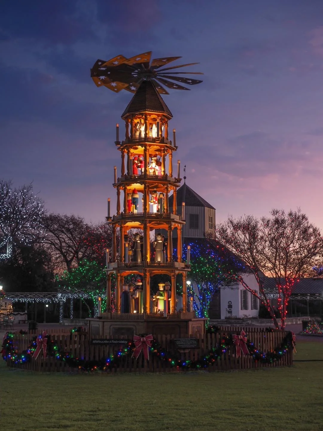A multi-tiered Christmas carousel with figurines, decorated with holiday lights and bows, set outdoors with Christmas trees and a building in the background during dusk.