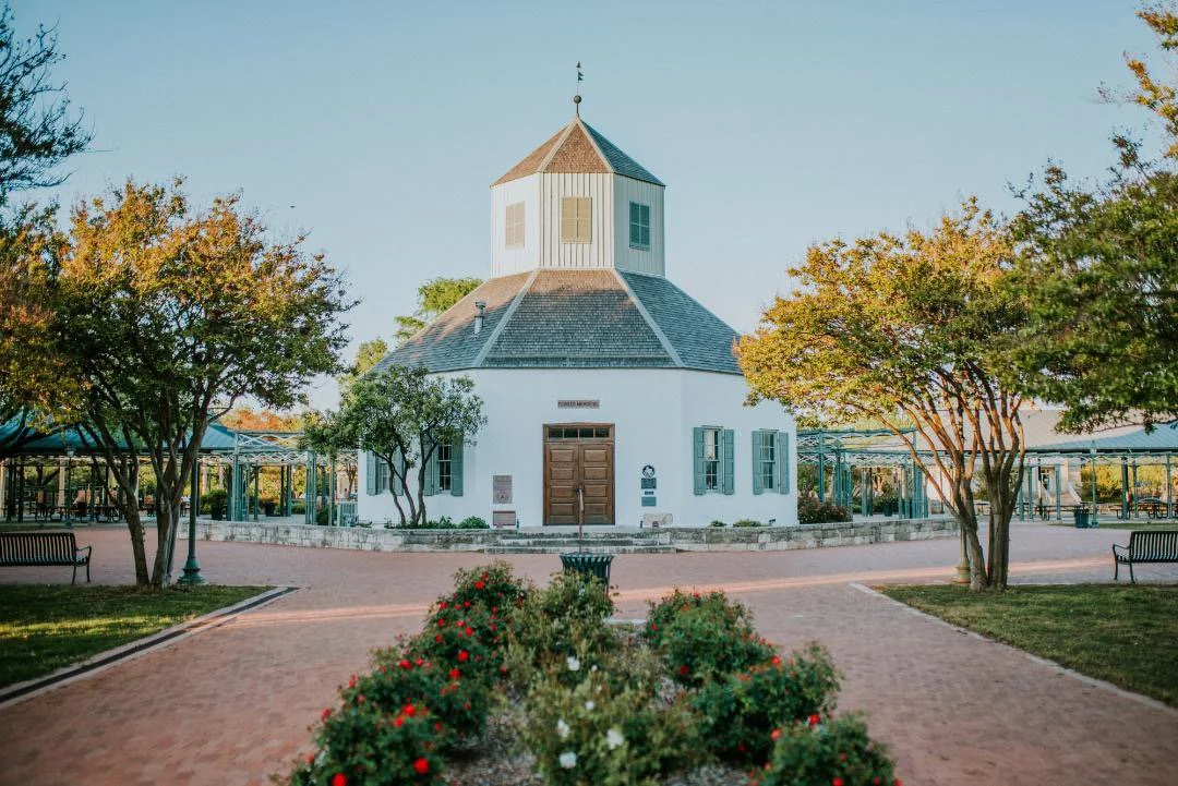 A white round building with a unique, tiered roof and blue shutters, surrounded by trees and landscaped with flowers and brick walkways.