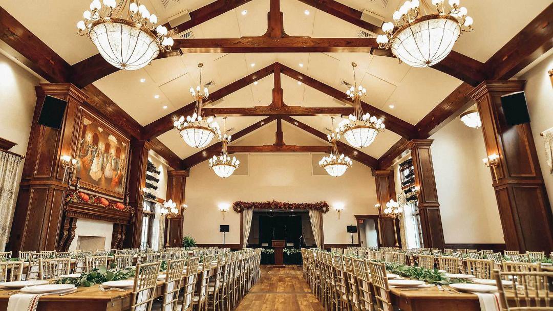 Elegant banquet hall decorated for an event, with long tables, gold chairs, chandeliers, paintings, and festive garlands.