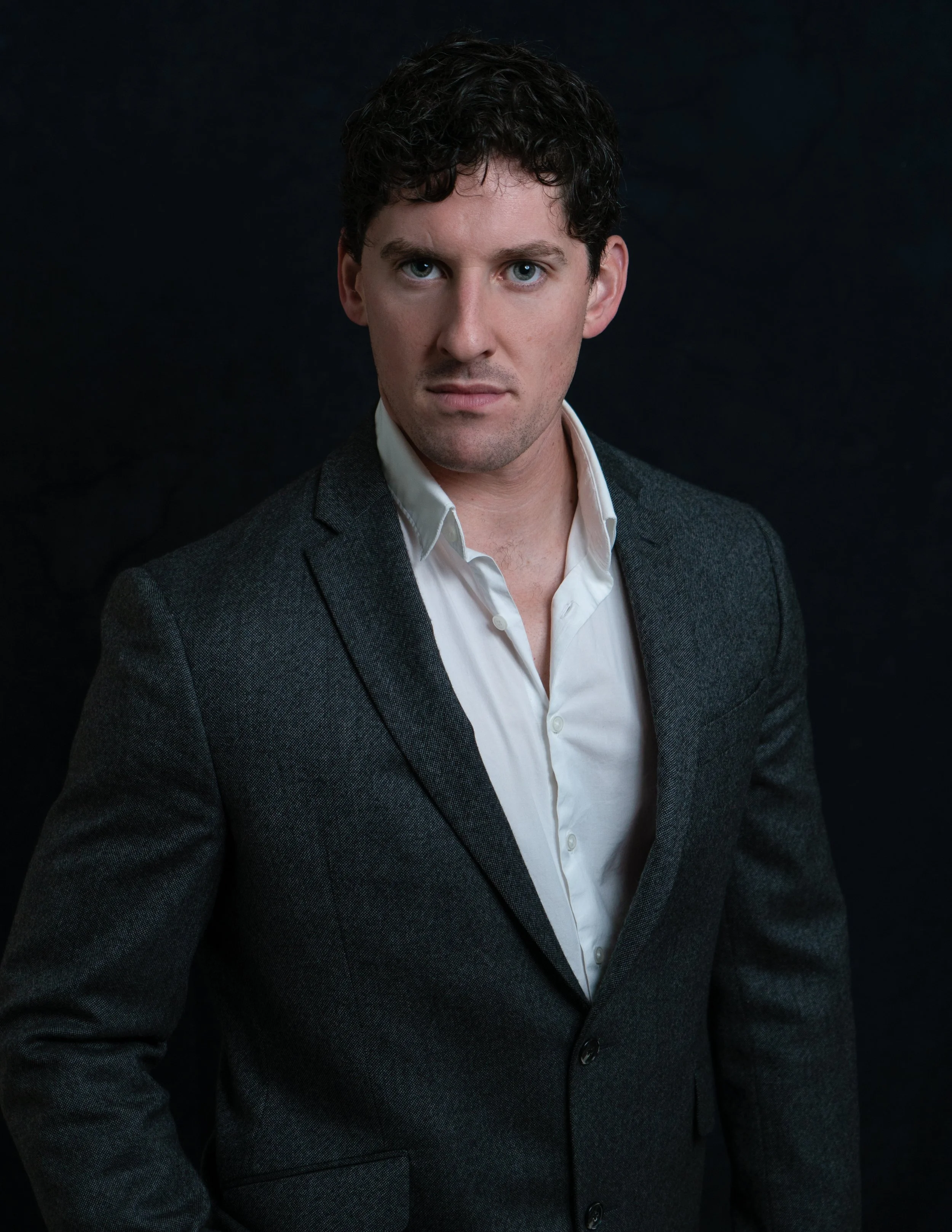 Man with dark curly hair wearing a white shirt and dark blazer against black background.