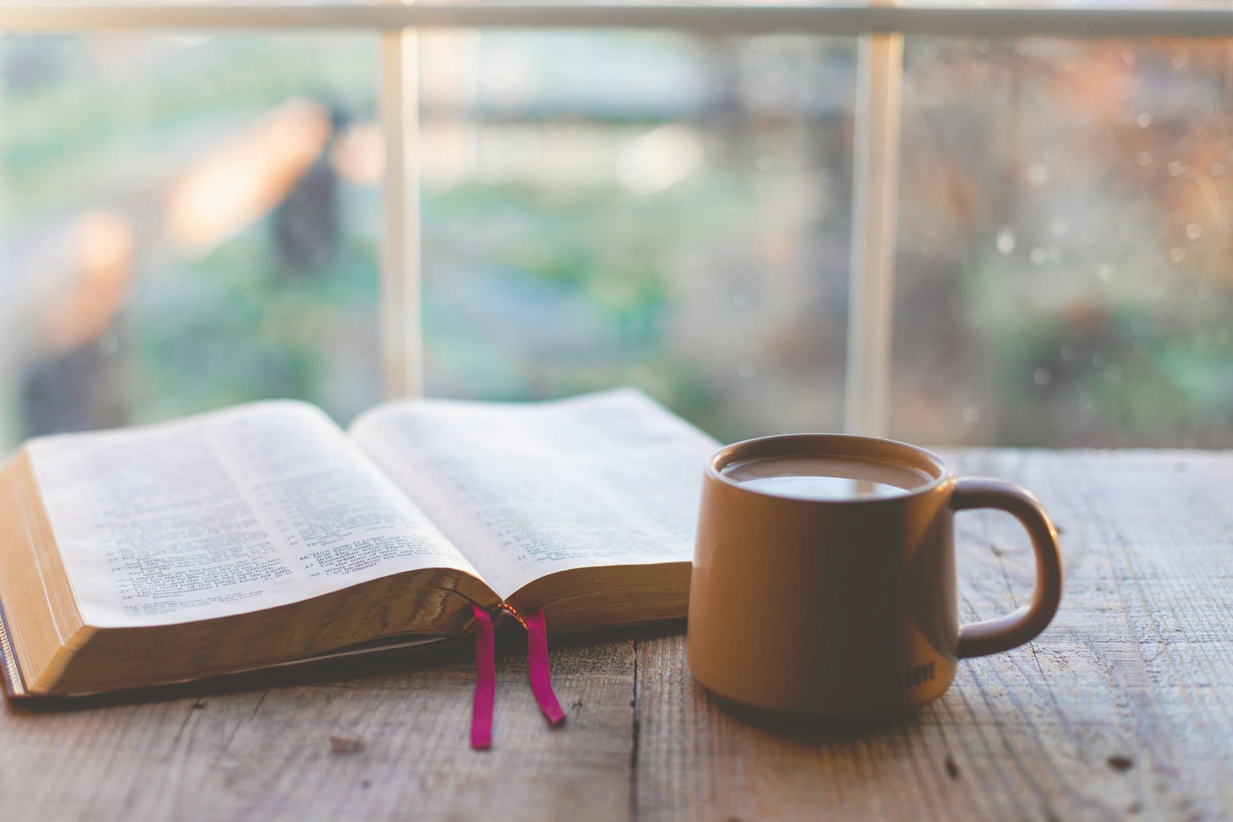 Open Bible and a mug of coffee on a wooden table near a window with blurred outdoor scenery.