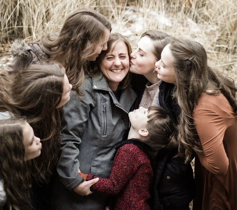 A woman and five girls hugging and kissing outdoors amid tall grass, showing affection and closeness.