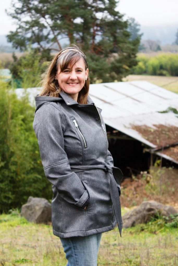 Smiling woman wearing a gray coat outdoors with green trees, rocks, and a corrugated metal shed in the background.