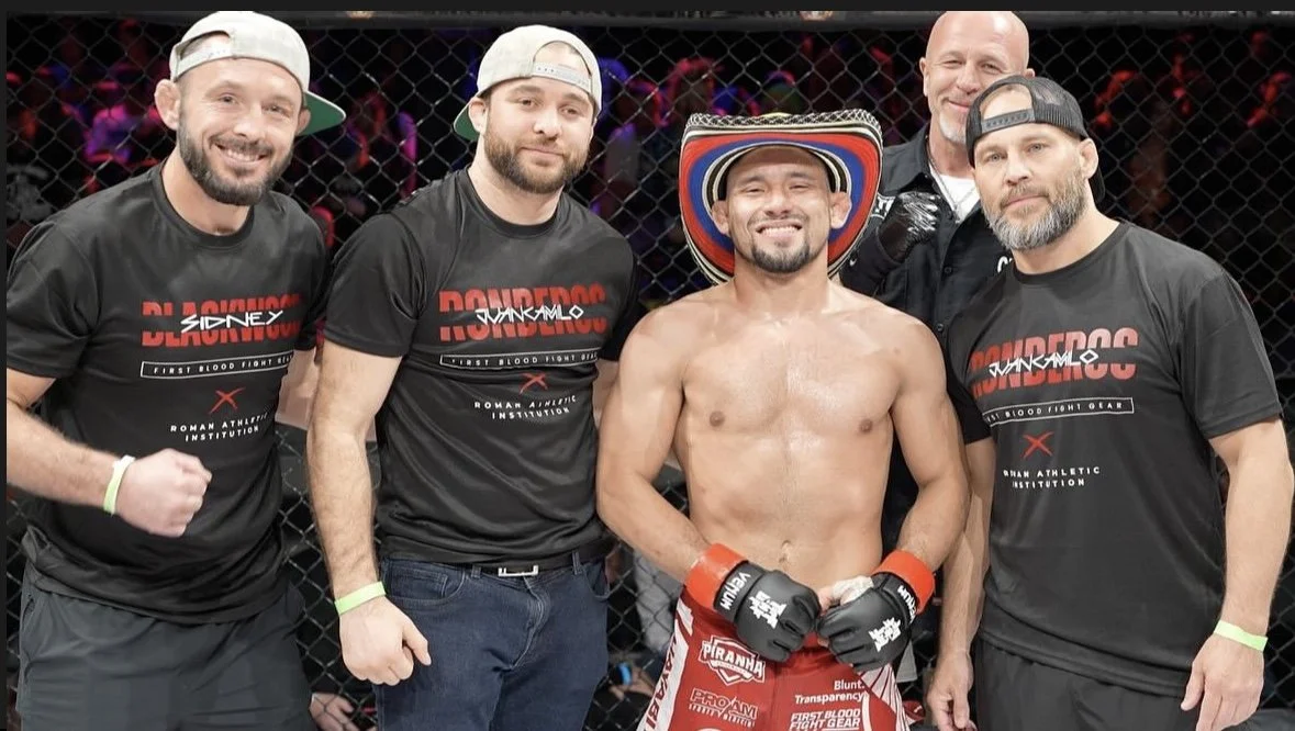Mixed martial arts fighter in red shorts and a sombrero hat, smiling with fists clenched, surrounded by four men in black shirts with red and white text, inside a cage.