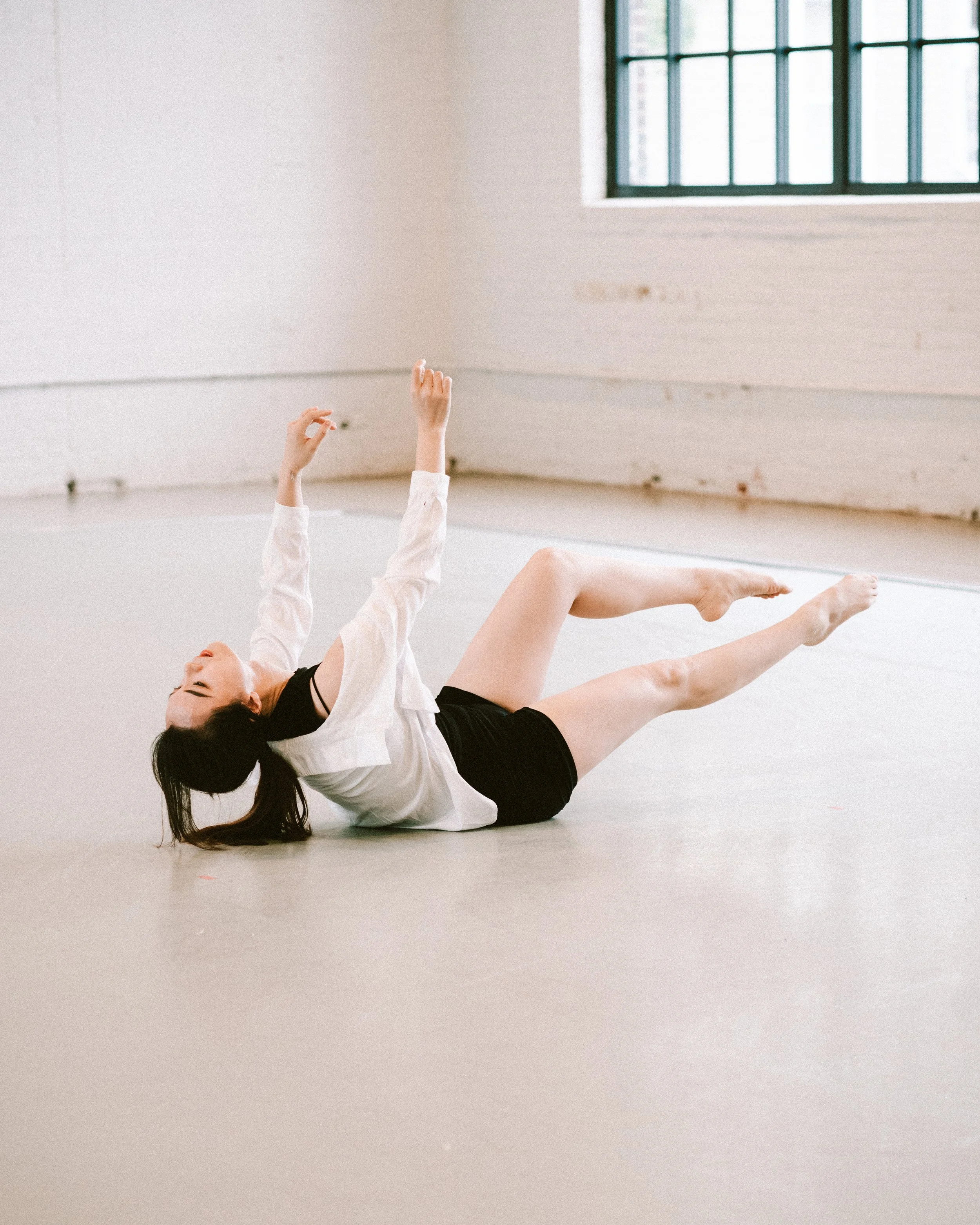 A person lying on their back on the floor with arms raised and legs bent, in a dance studio with a large window and white walls.