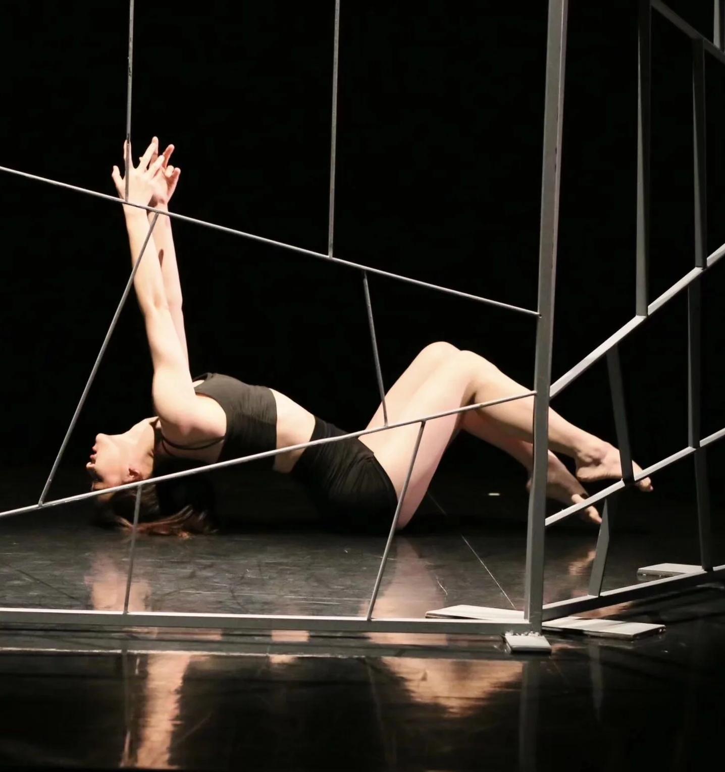 A woman lying on the floor inside a metal frame structure, with her head resting on the ground, and her arms extended upward, grasping the frame.