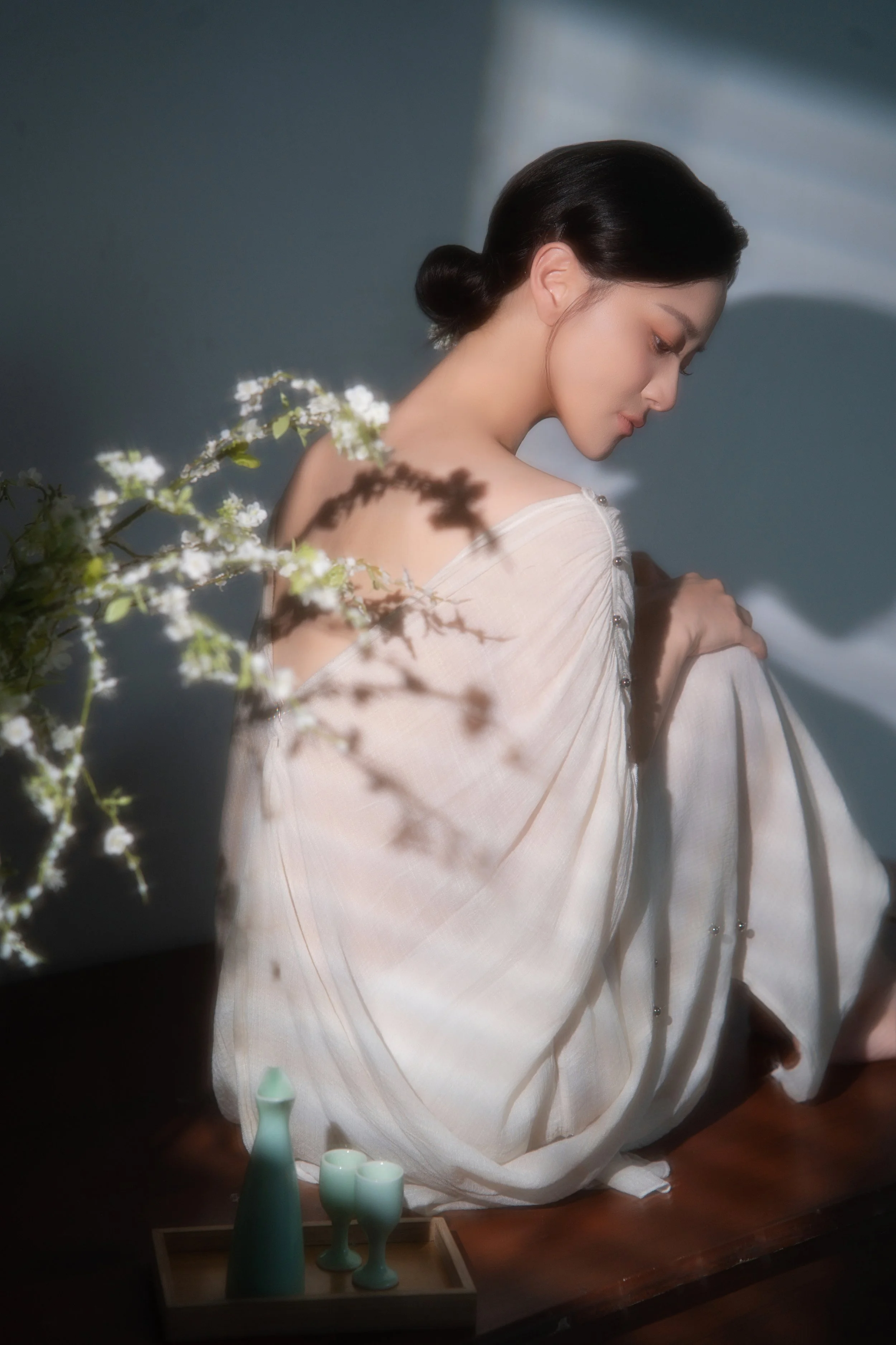 A woman with dark hair styled in a low bun, wearing a light-colored dress with pearl-like embellishments on the shoulder, is sitting with her head tilted down and eyes closed. There are white flowers in the foreground and a set of green vases on a tray in front of her. The background is plain with soft lighting creating shadowed areas.