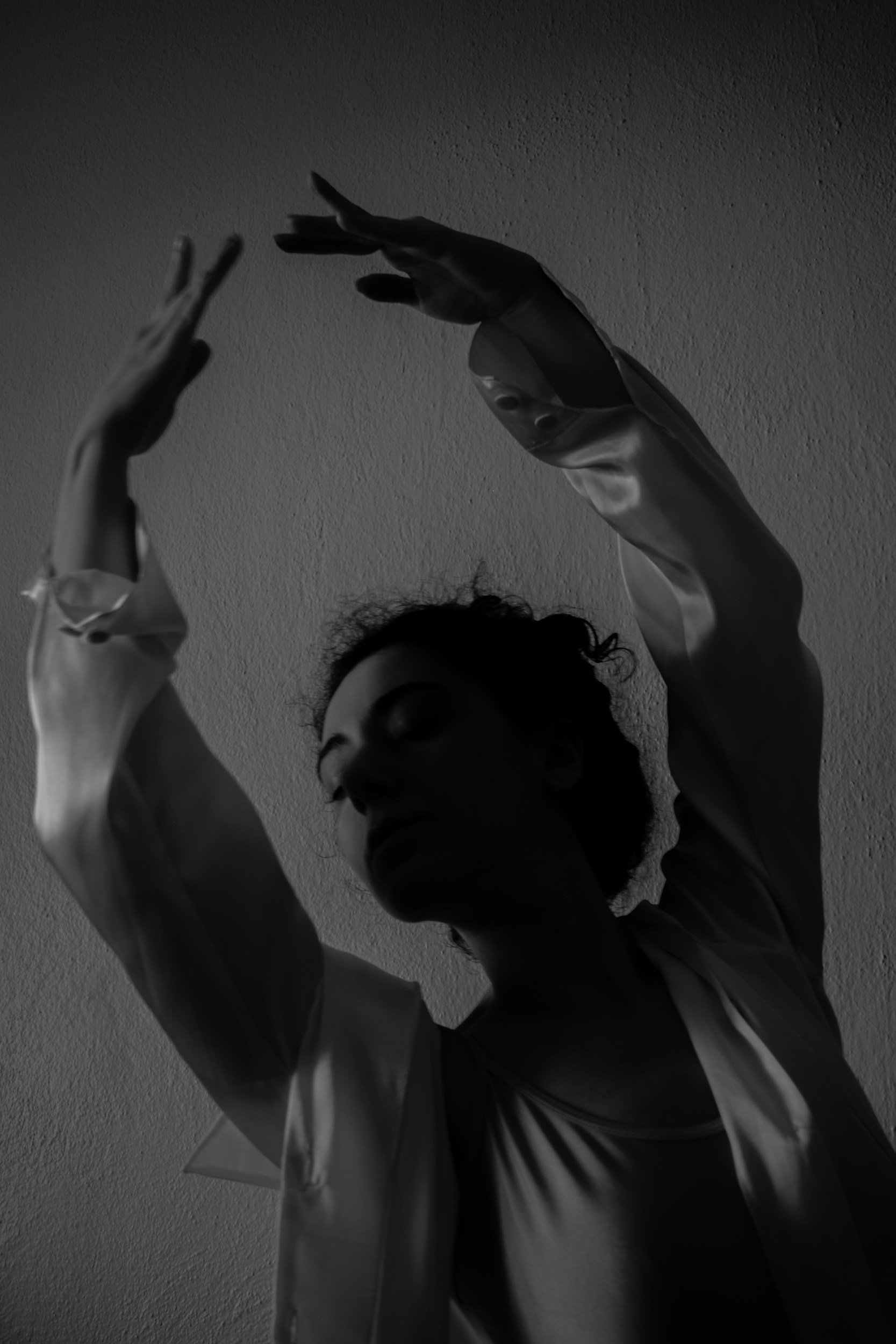 A black and white photo of a person with curly hair, arms raised, with light reflecting off metallic gloves, against a textured wall.