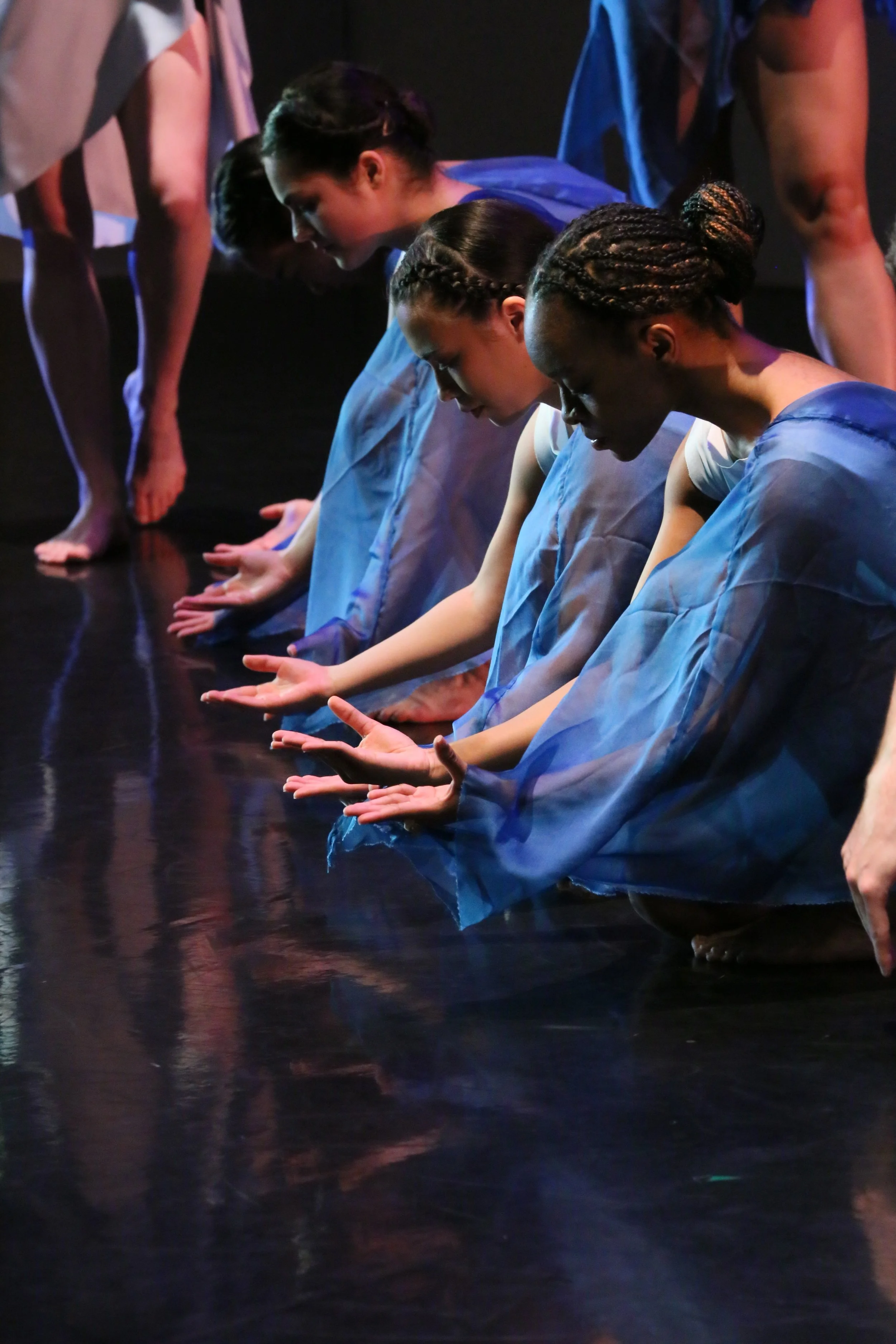 Group of dancers in blue costumes kneeling on stage with their hands extended forward in a theatrical performance.