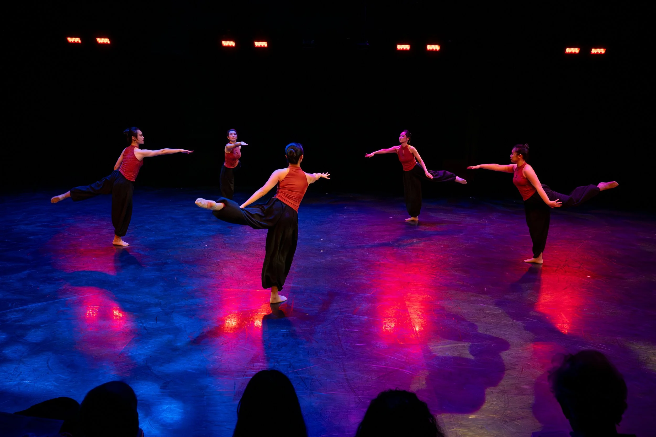 Five dancers practicing ballet on stage with colorful lighting, wearing red tank tops and black pants.