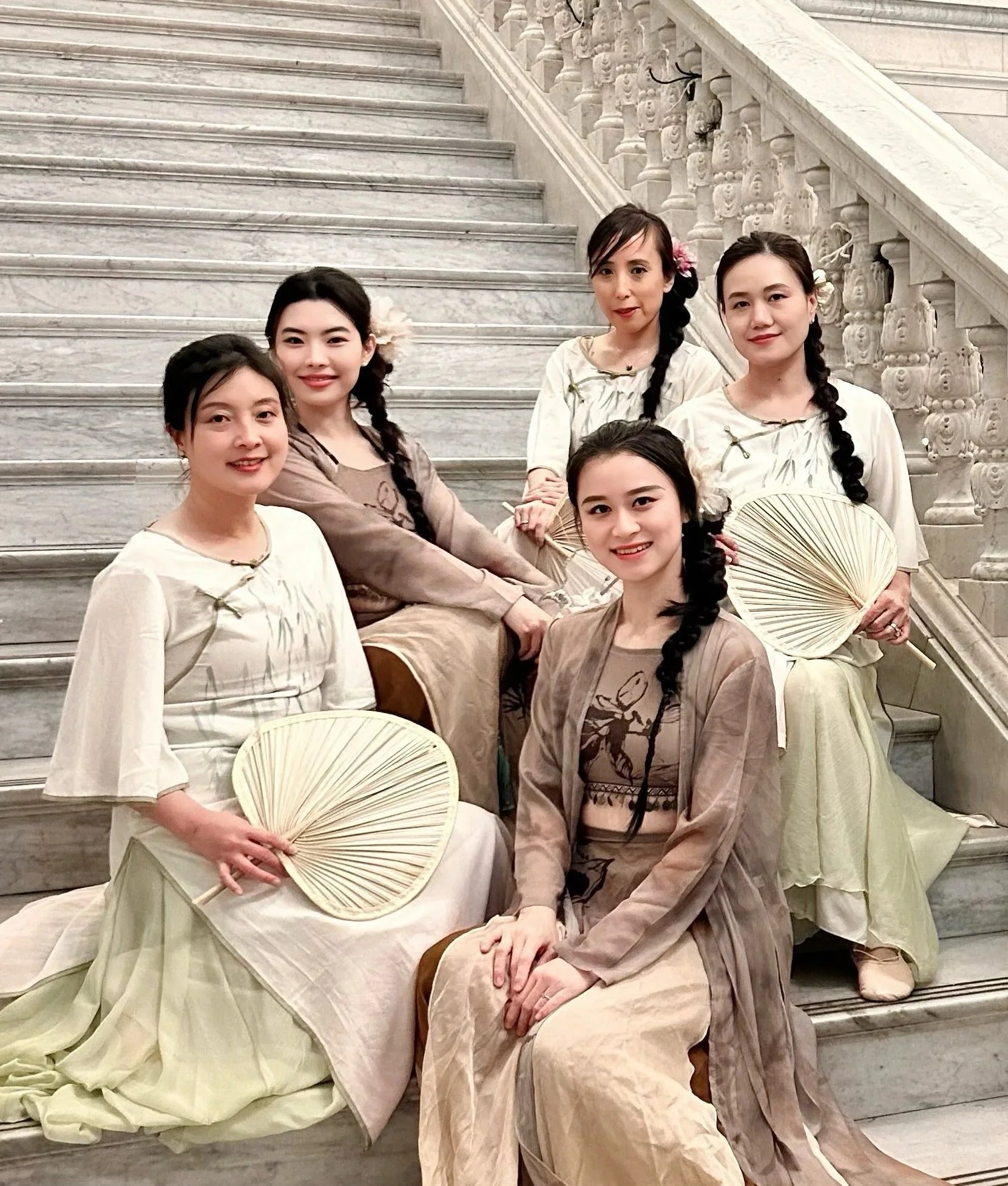 Five women sitting on a marble staircase, dressed in traditional Asian attire, holding fans, with intricate marble railings in the background.