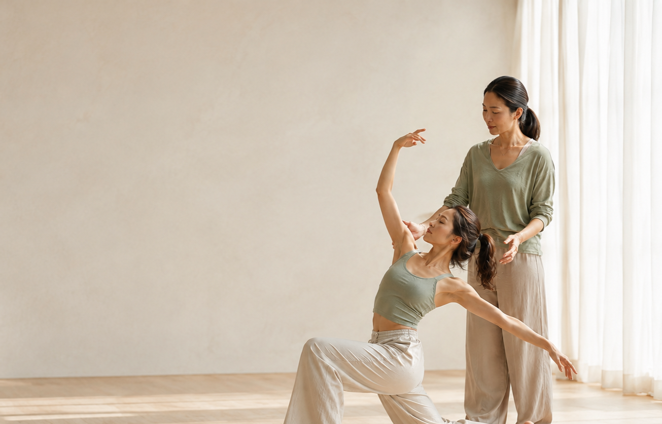 Two women practicing dance in a bright room with wooden floors and beige walls. One woman is sitting and stretching with her arms raised, while the other woman is standing and assisting.