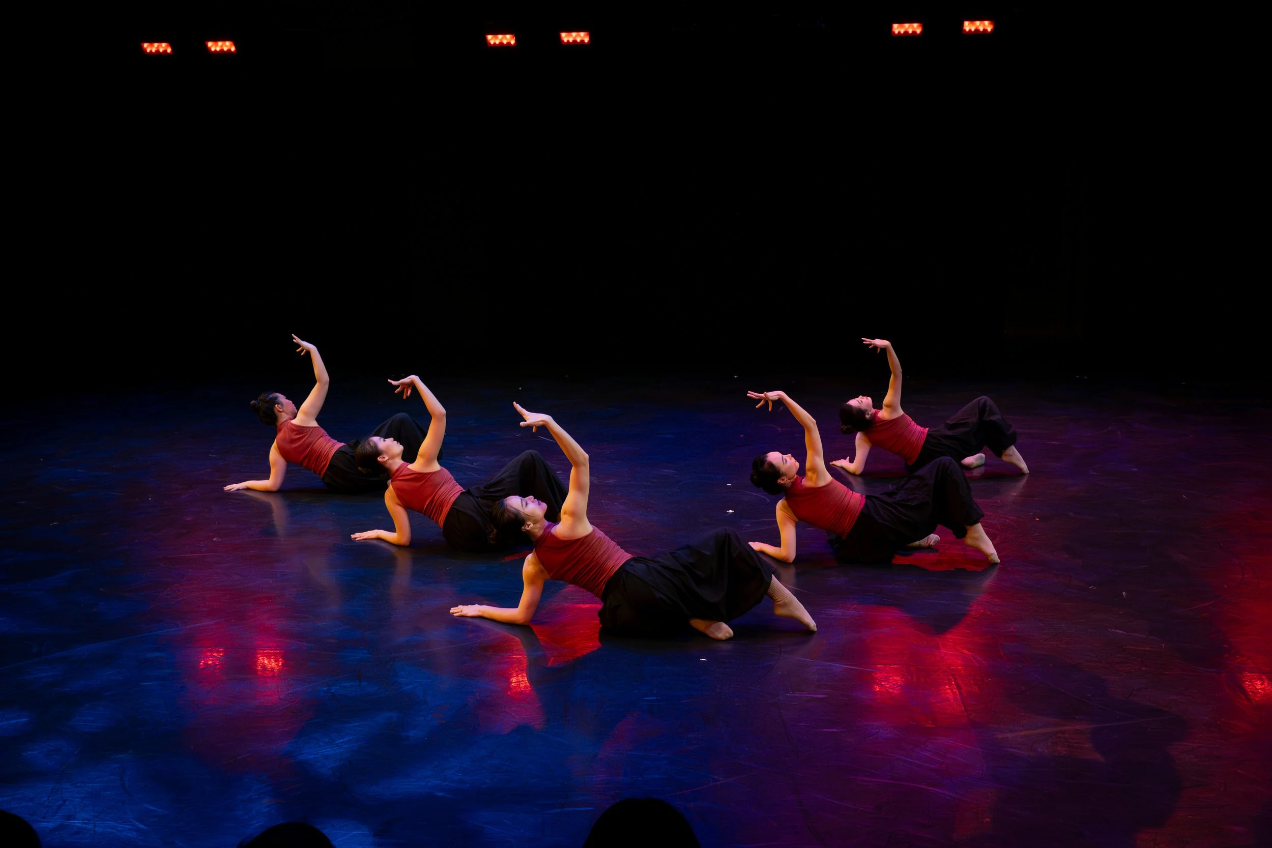 A dance performance with five women lying on the stage floor in matching red tank tops and black pants, with stage lights overhead creating a colorful lighting effect.