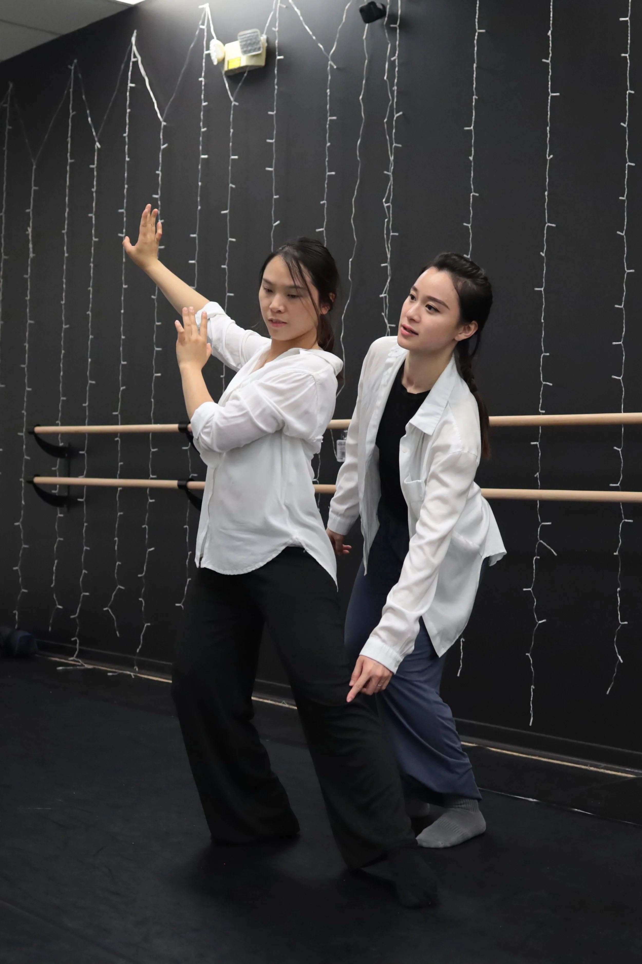 Two women in workout clothes practicing a dance move or pose in a dance studio with black floors, a black wall decorated with white string lights, and ballet barres along the wall.