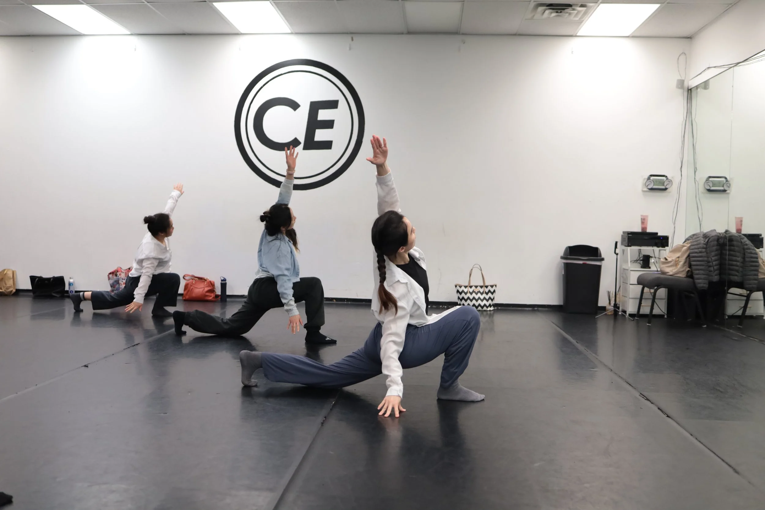 Four women participating in a yoga class in a studio, performing a pose with one knee and one hand on the floor, the other arm raised and looking towards the ceiling, with a large logo that reads 'CE' on the wall behind them.