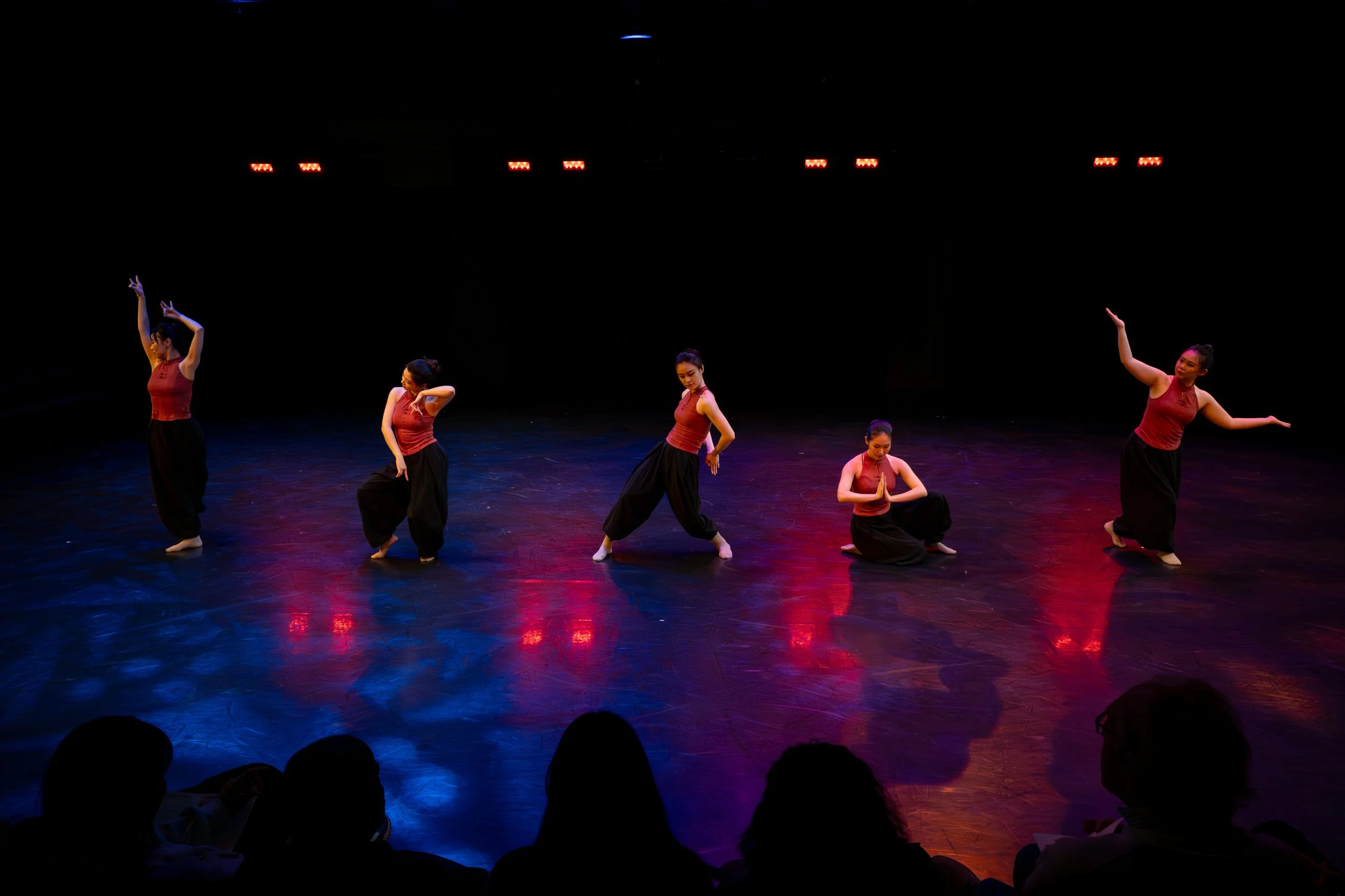 Five female dancers performing on a dimly lit stage with colorful reflections, wearing red tops and black pants.