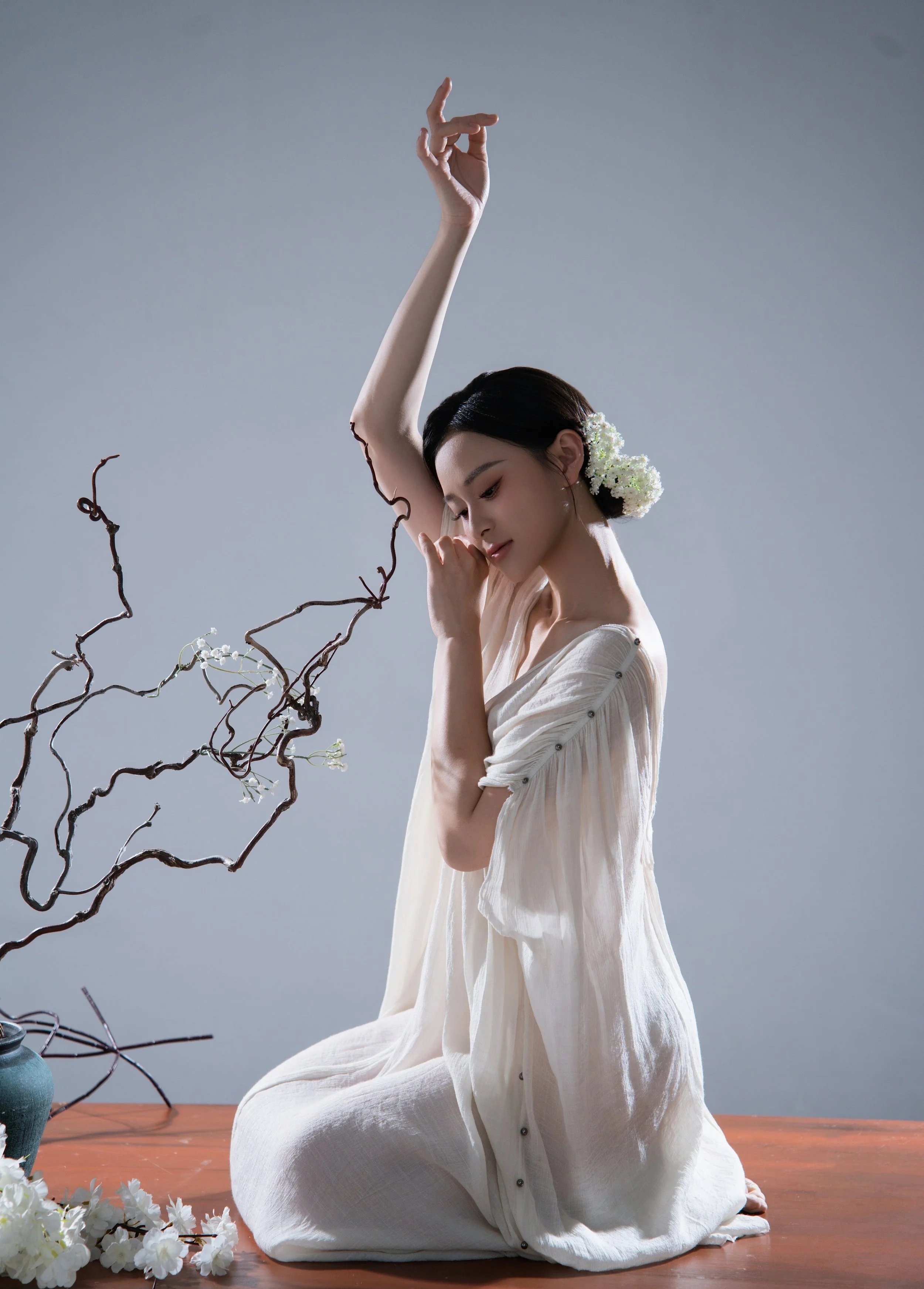 A woman with black hair, flowers in her hair, and wearing a light-colored dress kneeling on a wooden surface, raising one arm gracefully with flowers and twisted branches nearby.