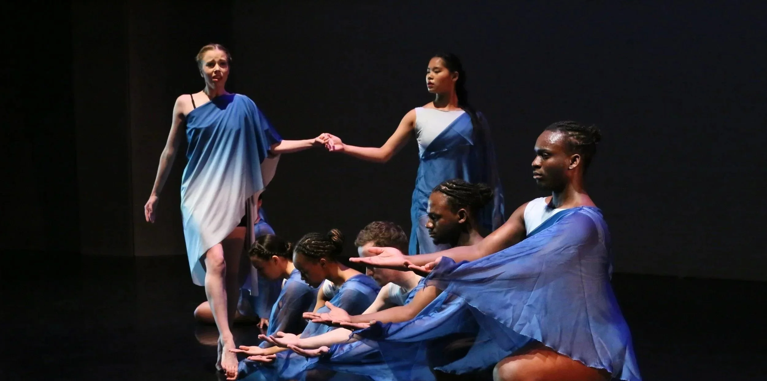 Diverse group of women in blue and white costumes participating in a theatrical or dance performance on stage, with some kneeling and others standing, holding hands and performing choreographed movements.