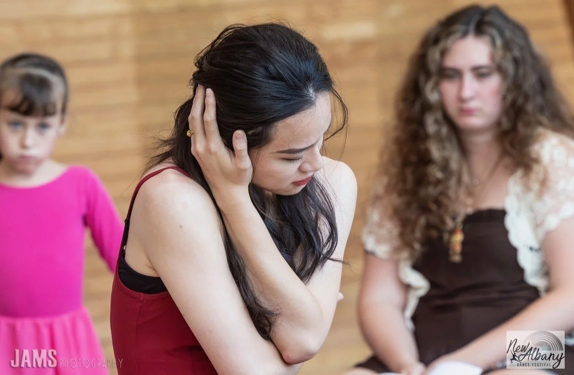 Three women are sitting close together in an emotional moment. The woman in the foreground with long dark hair is touching her head and appears upset. The woman on the right has curly hair and is looking down, also appearing emotional. The woman in the background is wearing a pink dress and is looking at the two women.