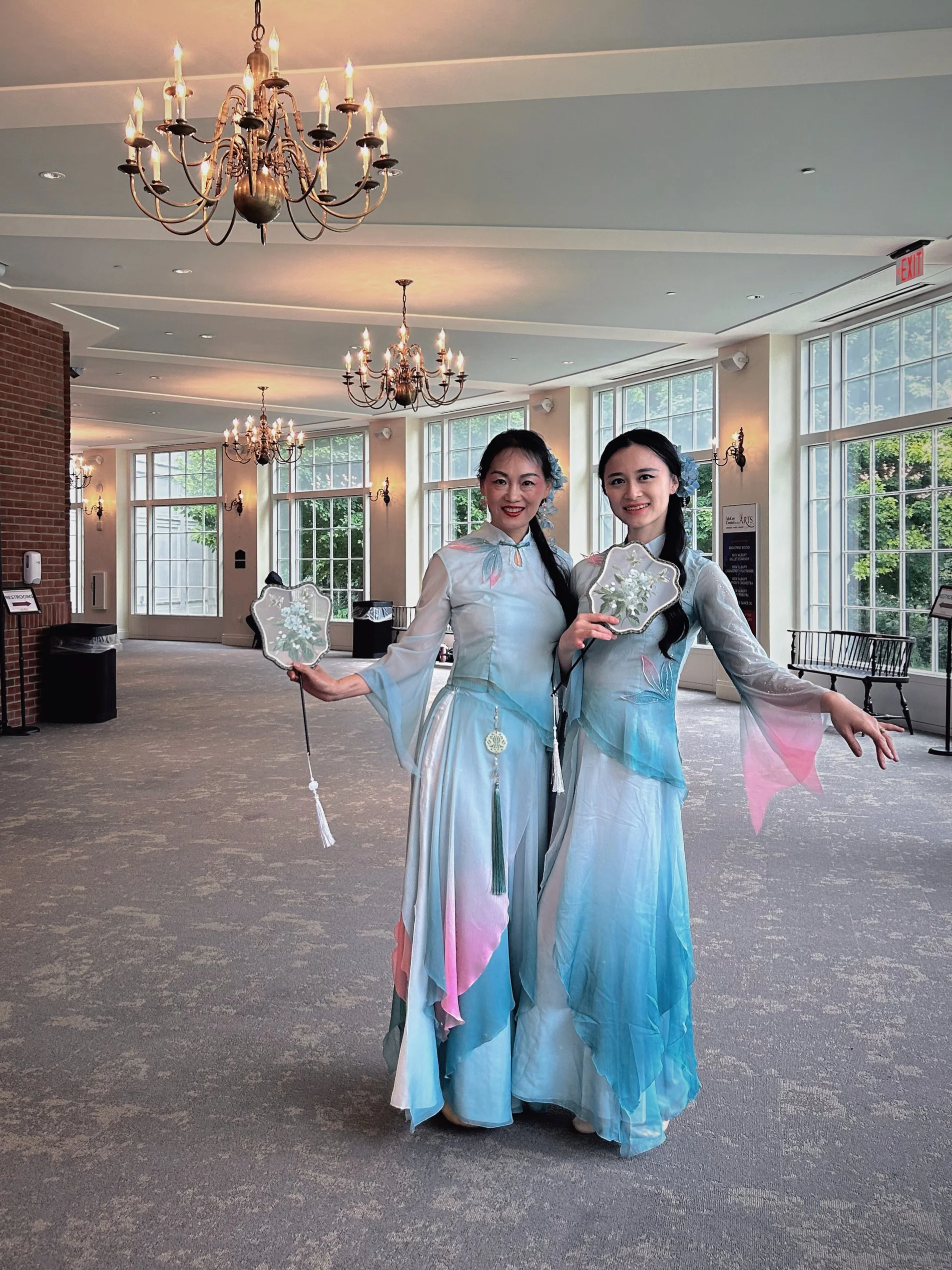 Two women wearing traditional Vietnamese áo dài dresses, holding fans, standing inside a bright room with chandeliers and large windows.