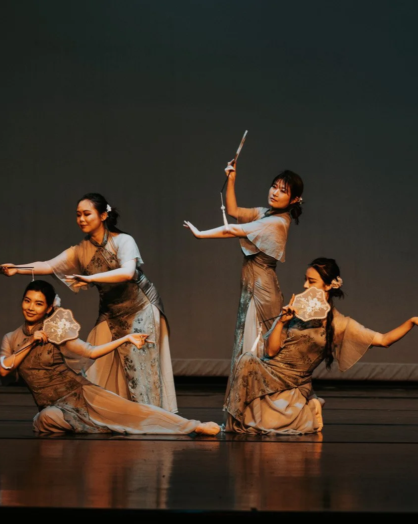 Four women in traditional Asian dance costumes performing on stage, holding fans and props.