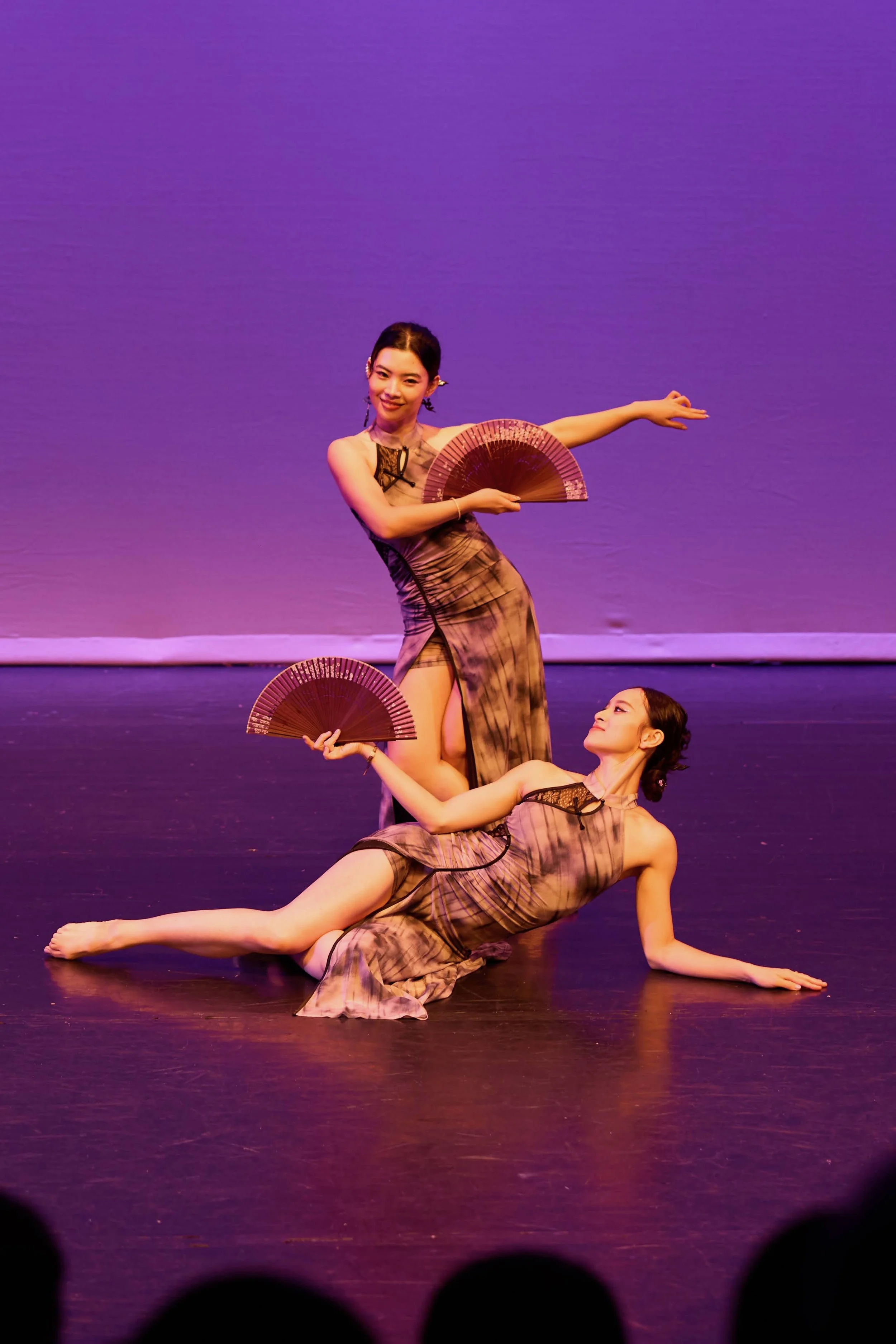 Two female ballet dancers performing on stage with purple background, wearing matching patterned costumes, holding fans, one lying on the floor and the other standing over her.