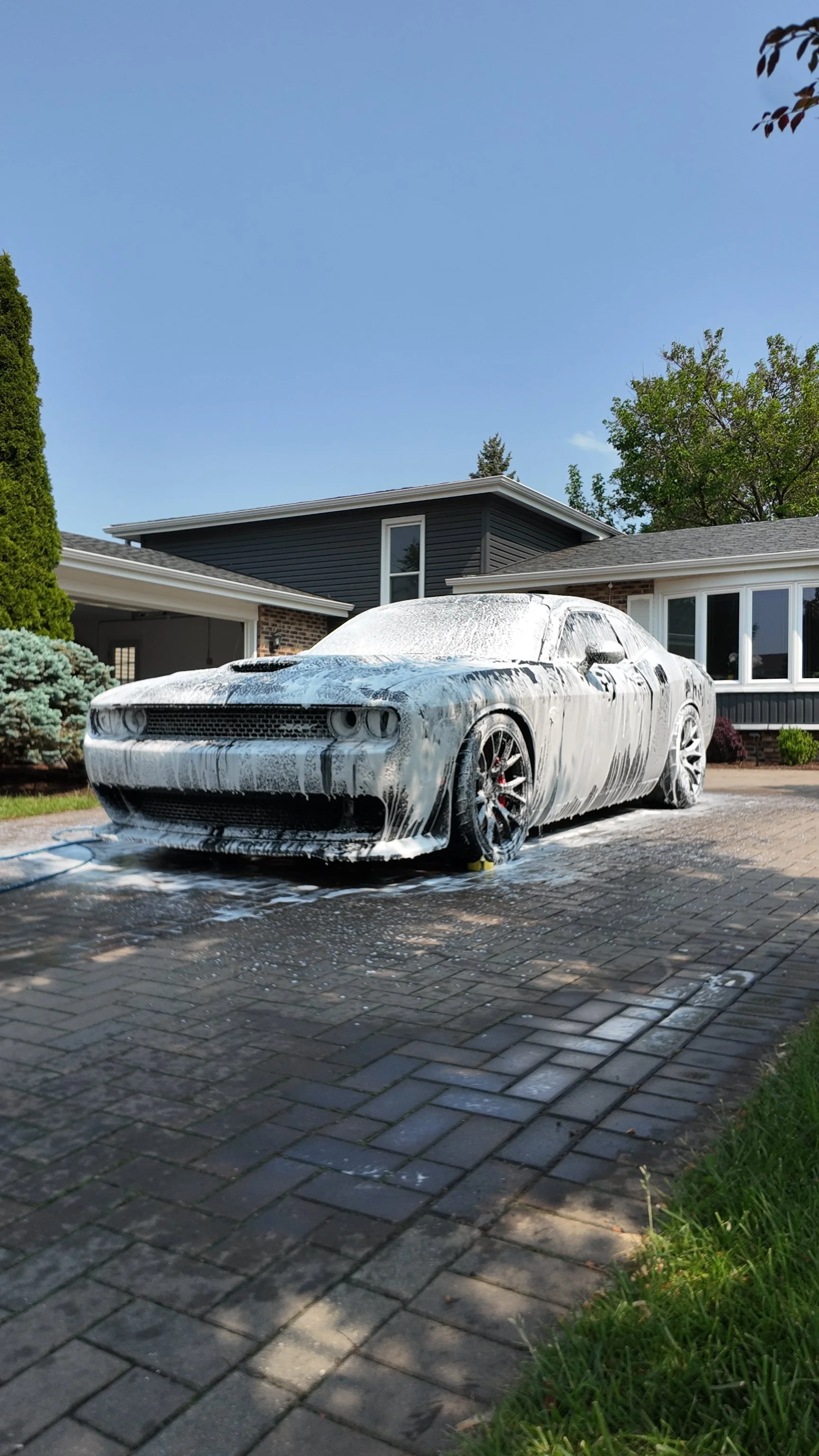 A silver sports car being washed with soap and foam in a driveway, next to a house with a yard and trees.