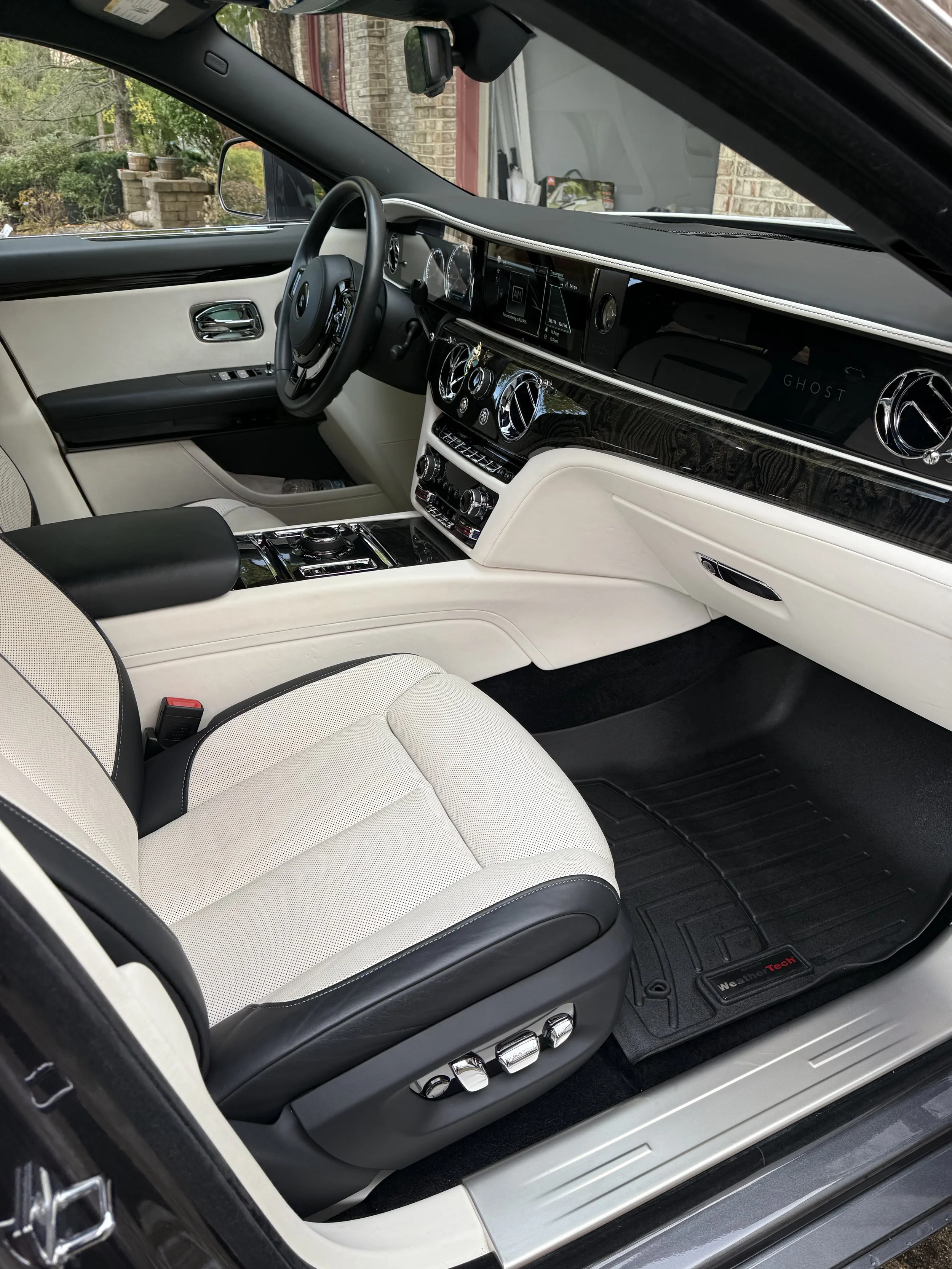 Interior of a luxury car showing black and cream seats, a black steering wheel, a sleek dashboard with multiple digital displays, and a gear shift area with controls. The car has a modern, upscale design.