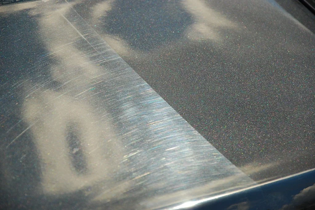 Close-up image of an automobile's hood showing a noticeable scratch and a duller, rough area with some specks of dirt or rust.