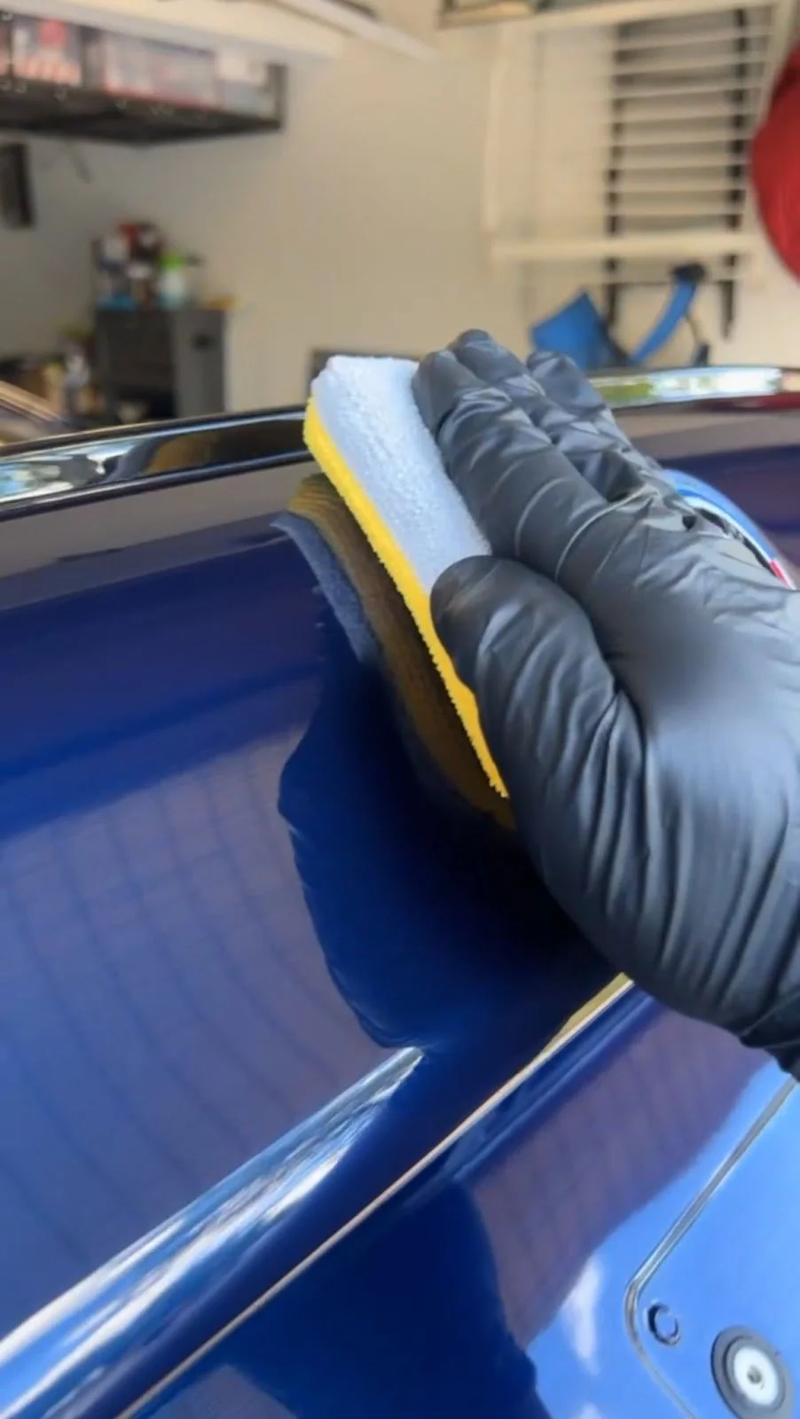 A person wearing a black glove using a yellow and white sponge to polish the surface of a blue car in a garage.