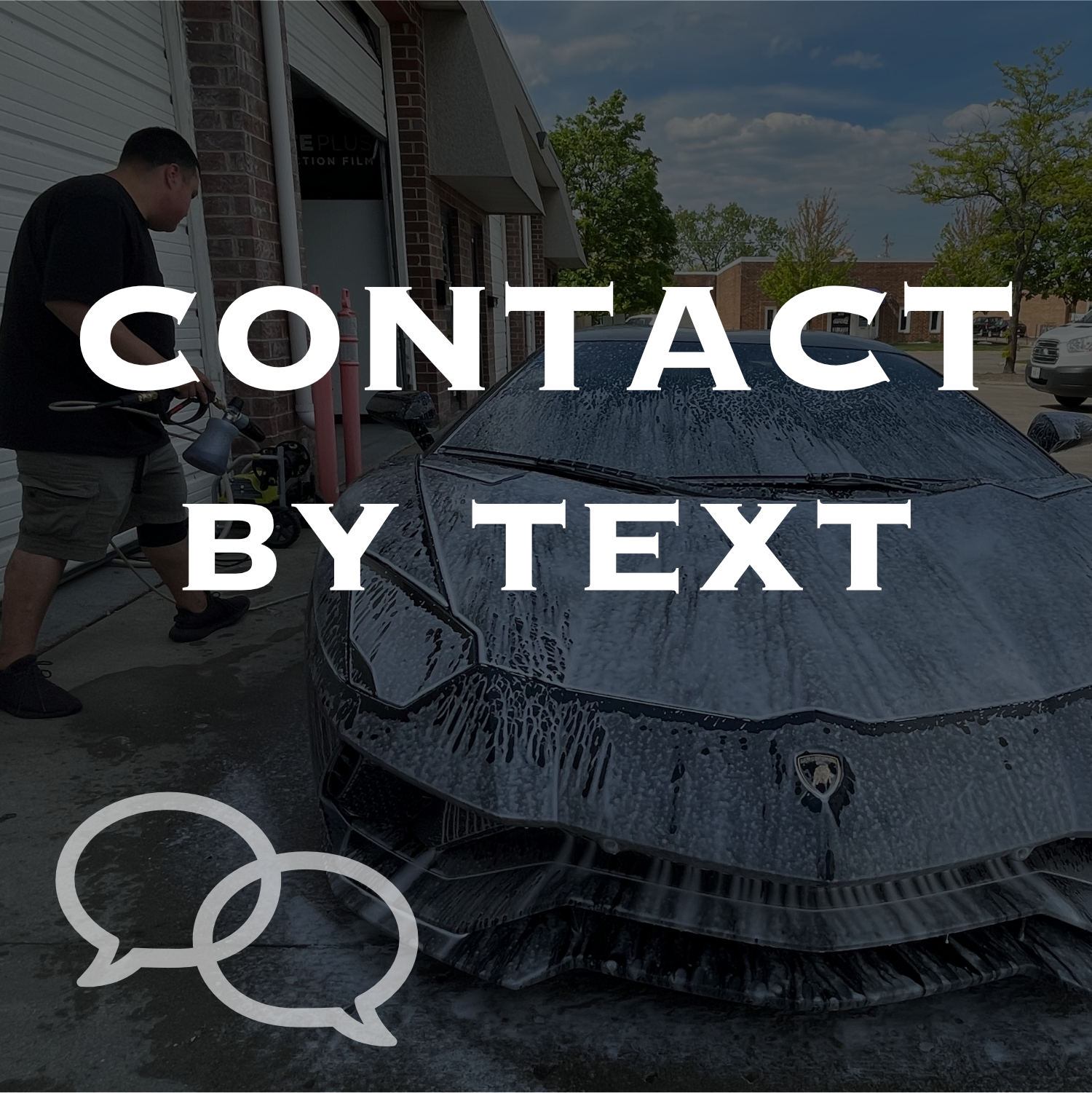 A black sports car, possibly a Lamborghini, covered in soap suds during a car wash outside a building, with a man washing the car and several other vehicles in the background.