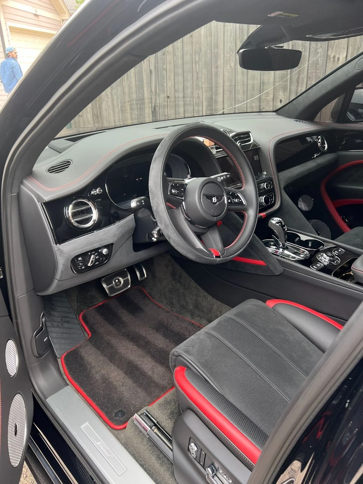 Interior of a luxury car featuring a leather and suede steering wheel with red stitching, digital dashboard, control buttons on the center console, and black and red upholstered seats.
