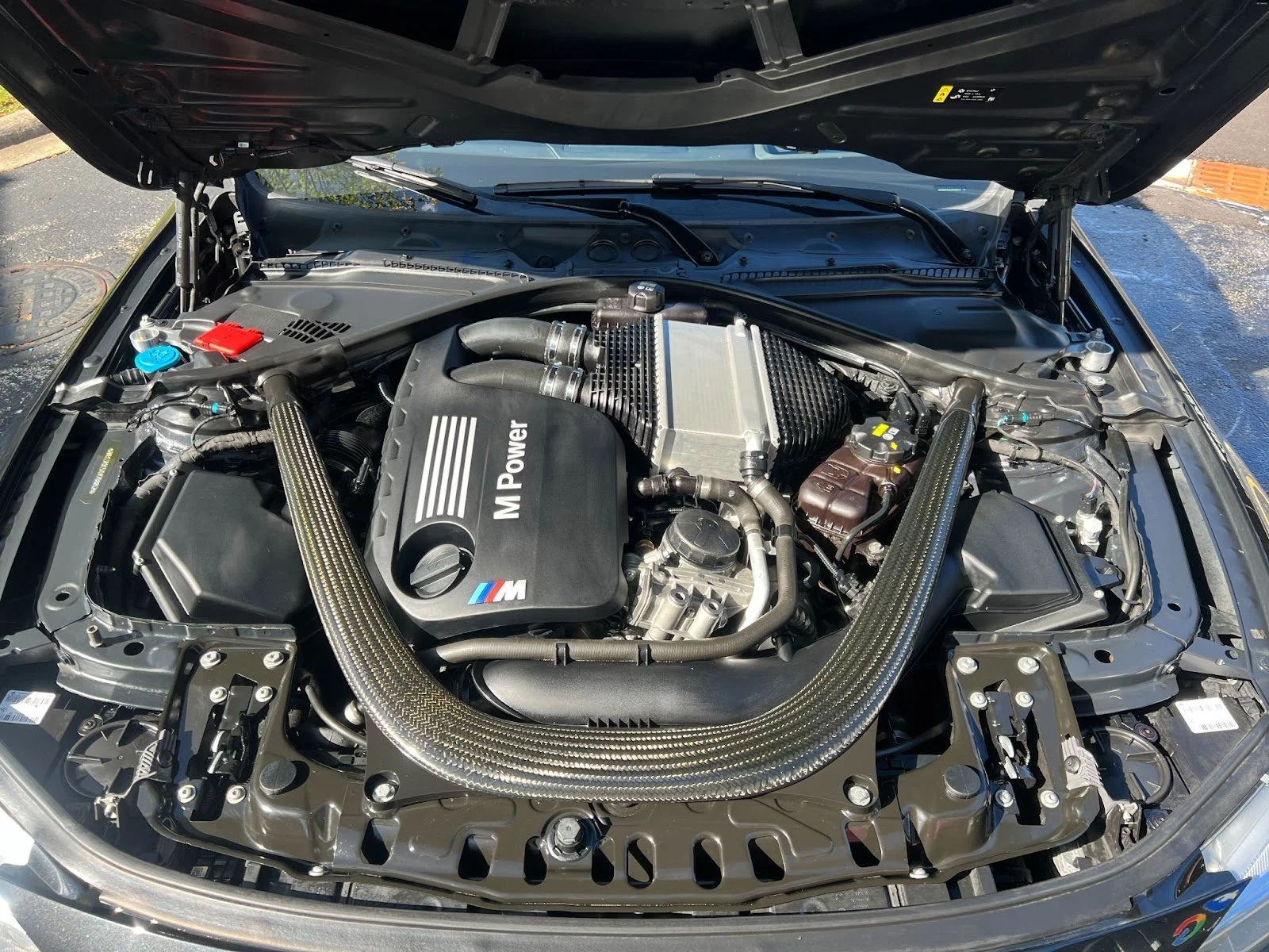 Car engine bay with BMW M Power engine, carbon fiber strut braces, and various engine components.
