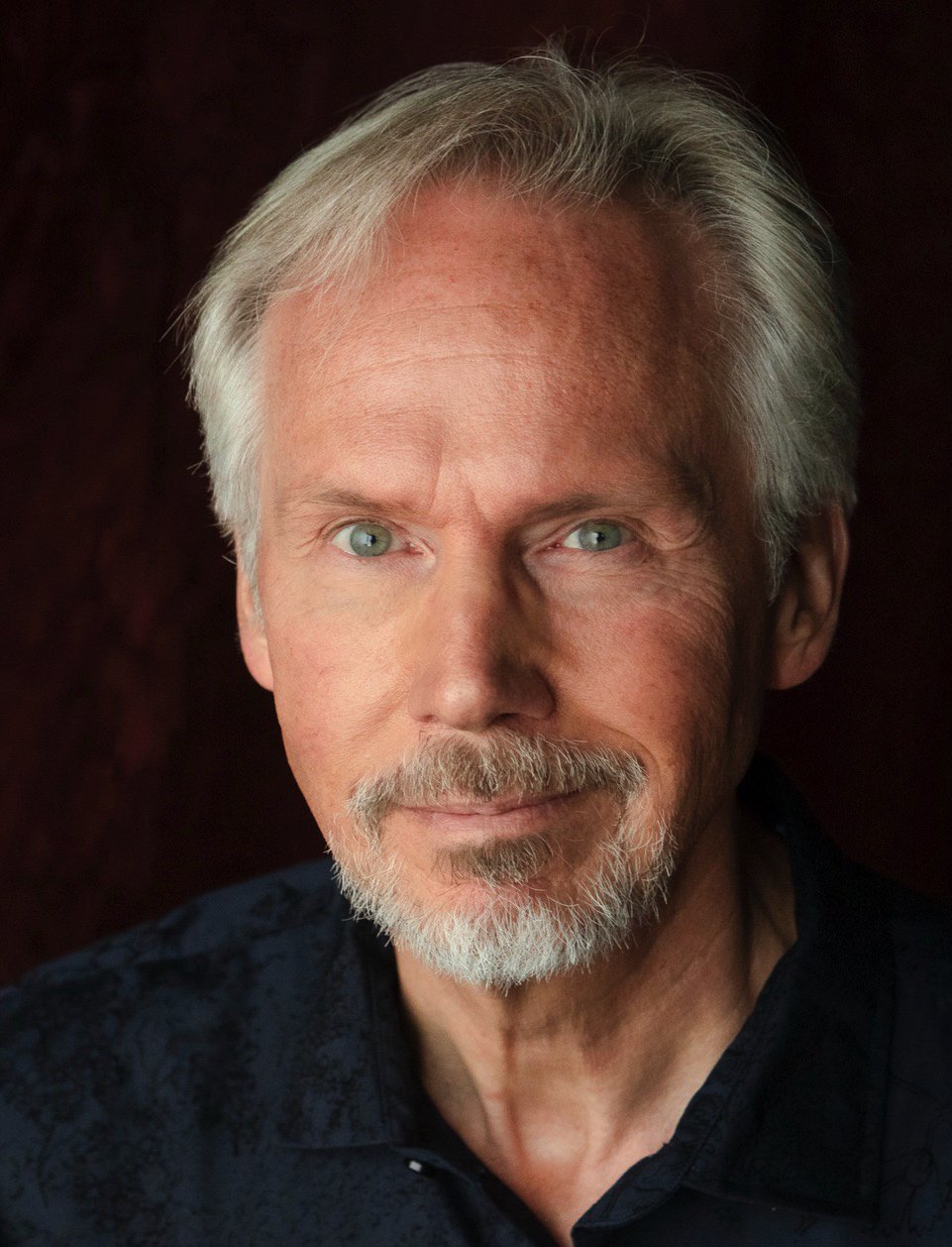 Close-up portrait of a senior man with white hair, a beard, piercing blue eyes, wearing a dark shirt, against a dark background.