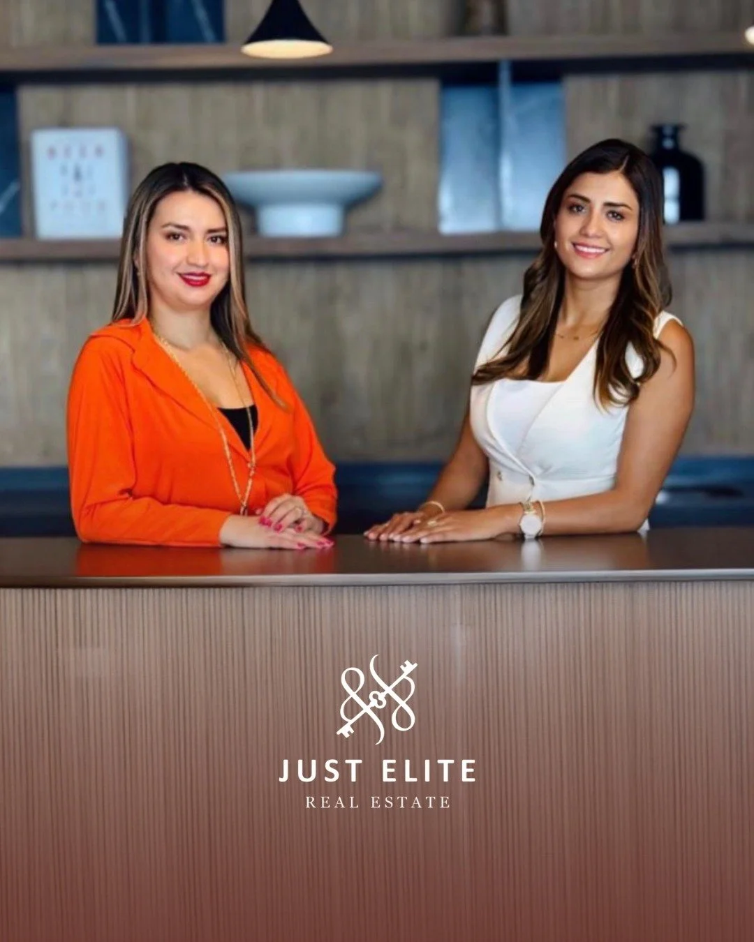 Two women standing behind a reception desk with 'Just Elite Real Estate' logo, smiling at the camera.