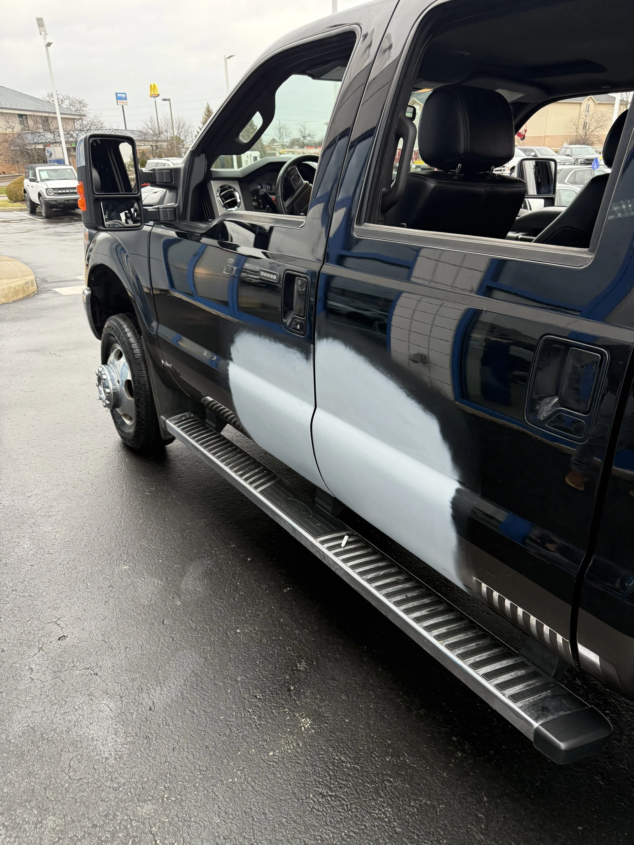 A black pickup truck with extended cab parked on a wet lot, showing part of the front and side view with a step bar below the door.