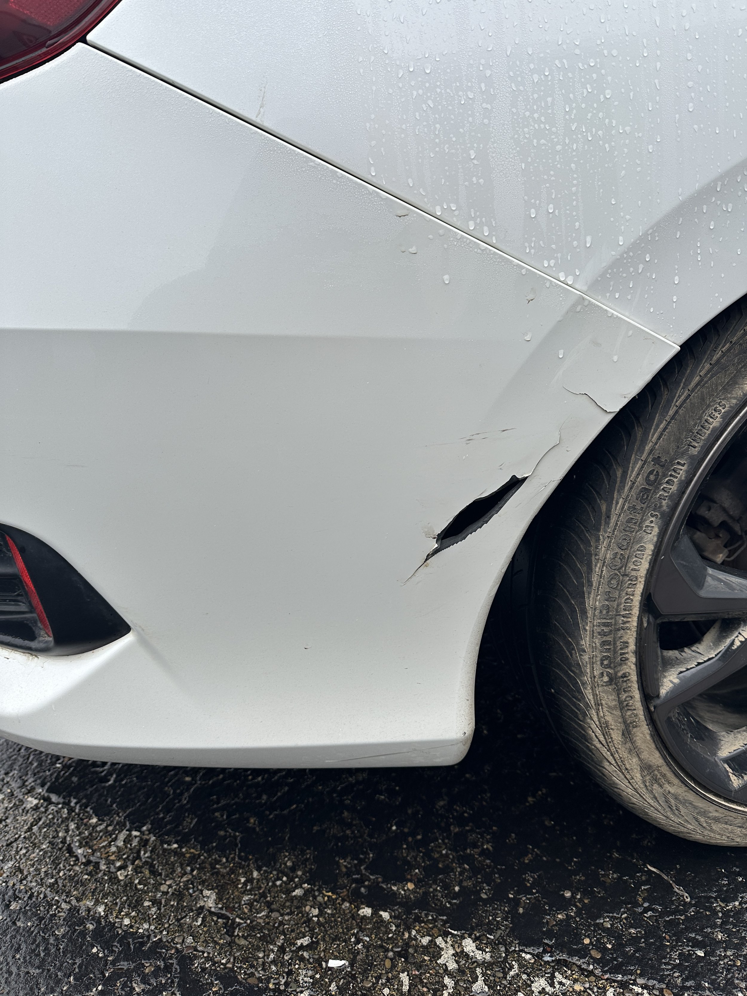 Close-up of a white car's front side with a large dent and scratches near the wheel well, and rain droplets on the surface.