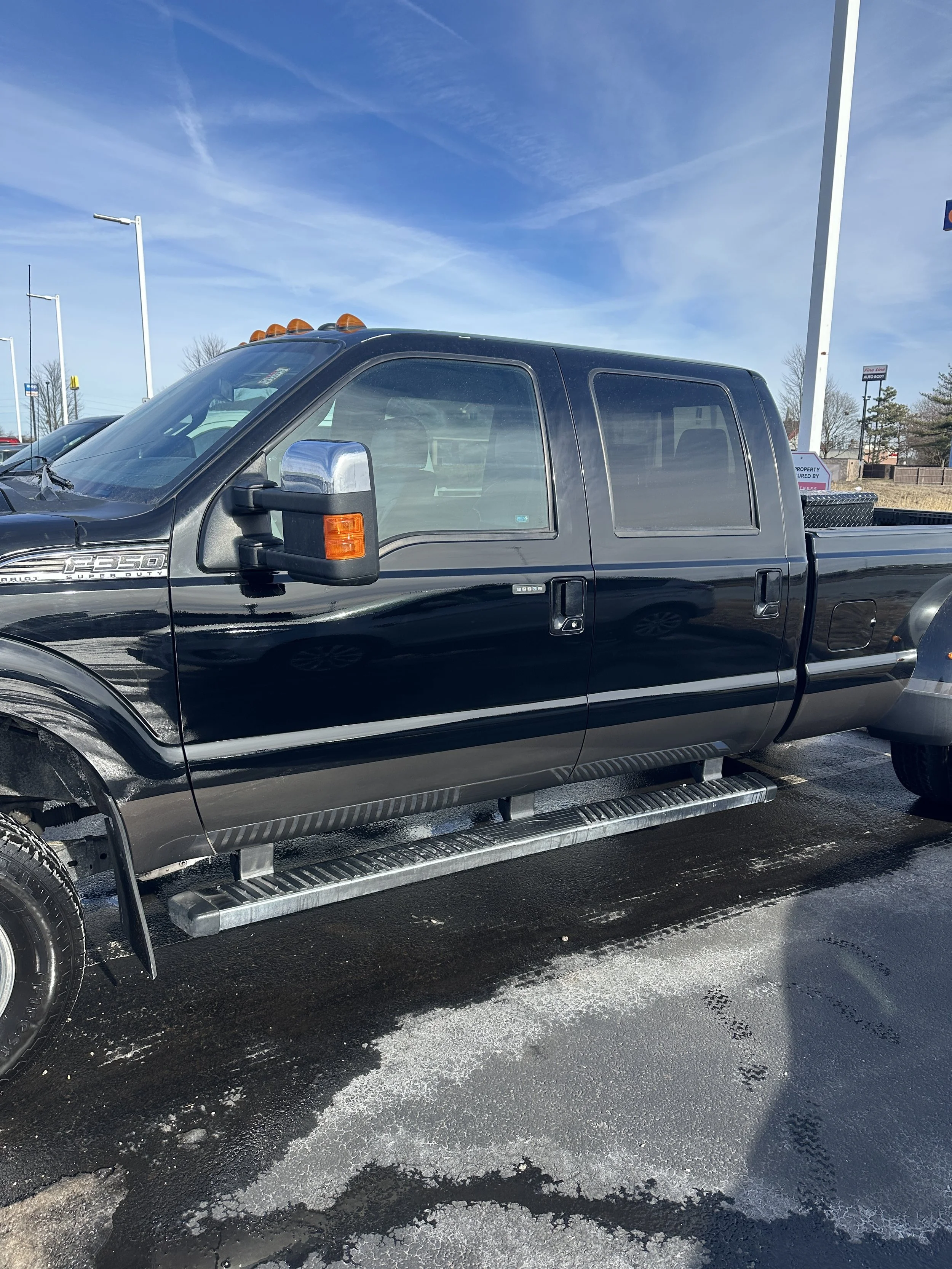 Black Ford F-250 pickup truck parked outdoors on a wet parking lot, with a blue sky and several light poles visible in the background.