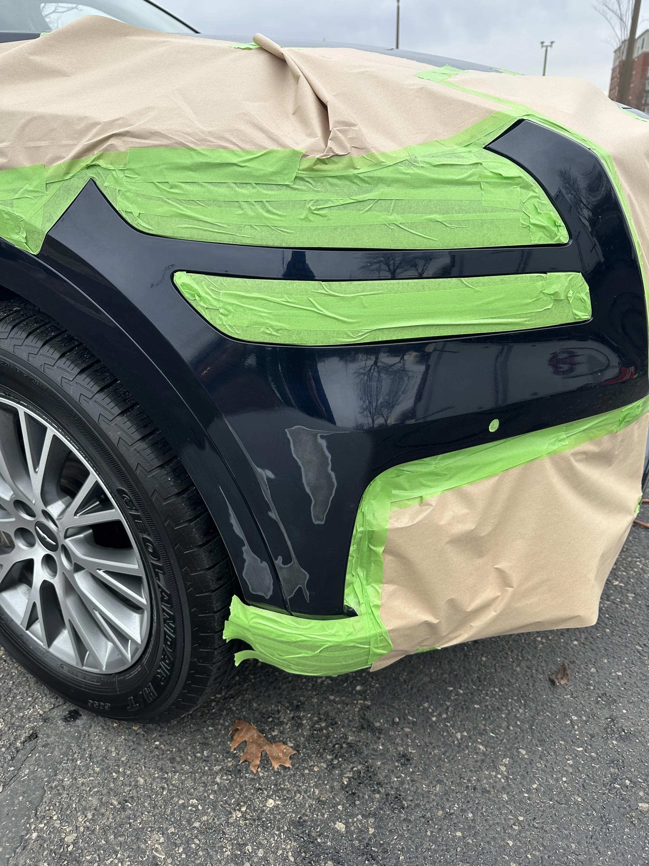 Close-up of a car undergoing bodywork repair, with green painter's tape and brown paper covering for painting or protection, parked on an asphalt surface with fallen leaves.