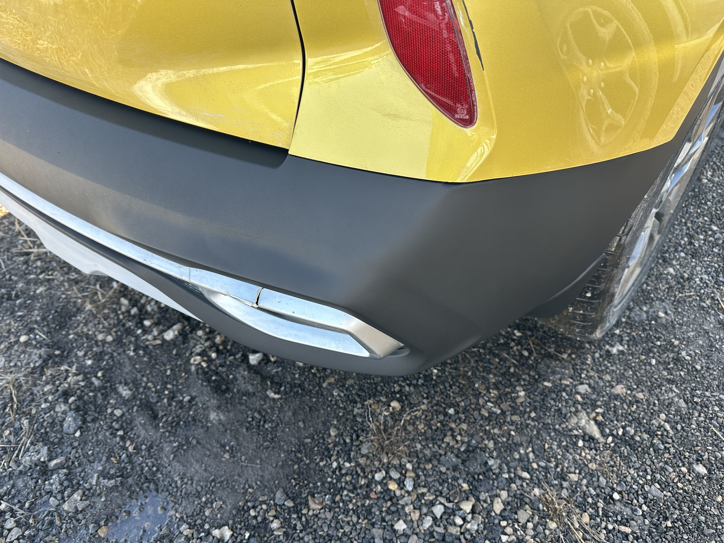 Close-up of the rear part of a yellow car showing the tail light, bumper, and part of the wheel on a gravel surface.