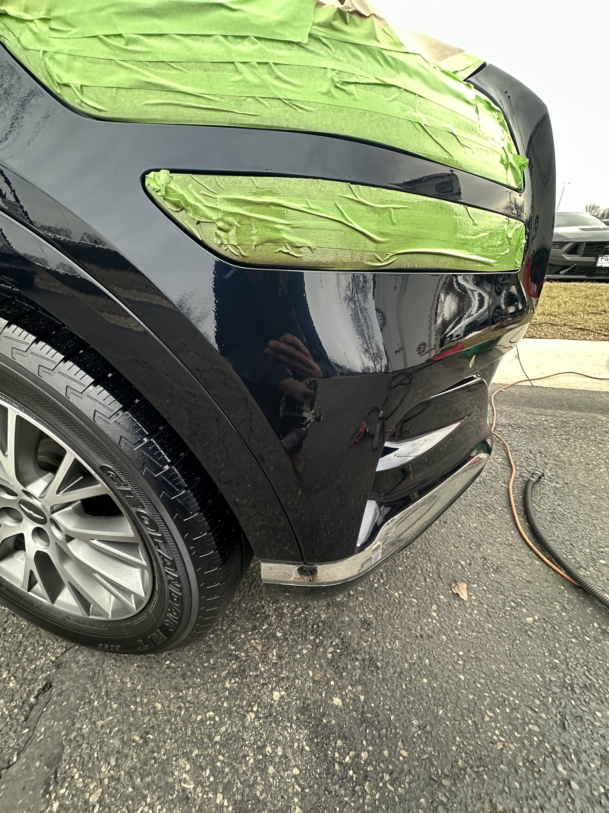 Close-up of the front right corner of a black car with green masking tape over the hood and fender, likely during a painting or repair process.