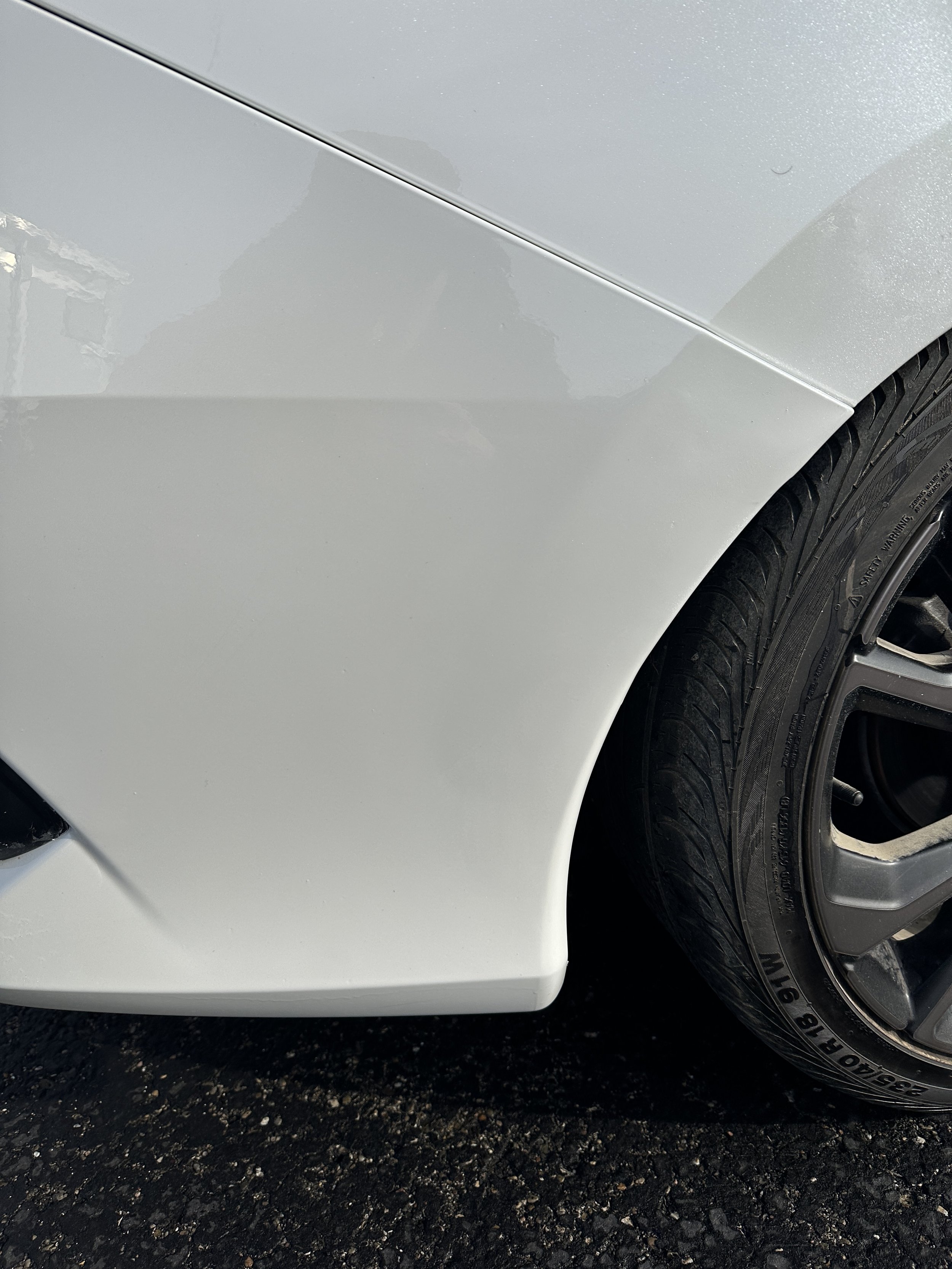 Close-up of a white car's front fender and tire on a black asphalt surface.