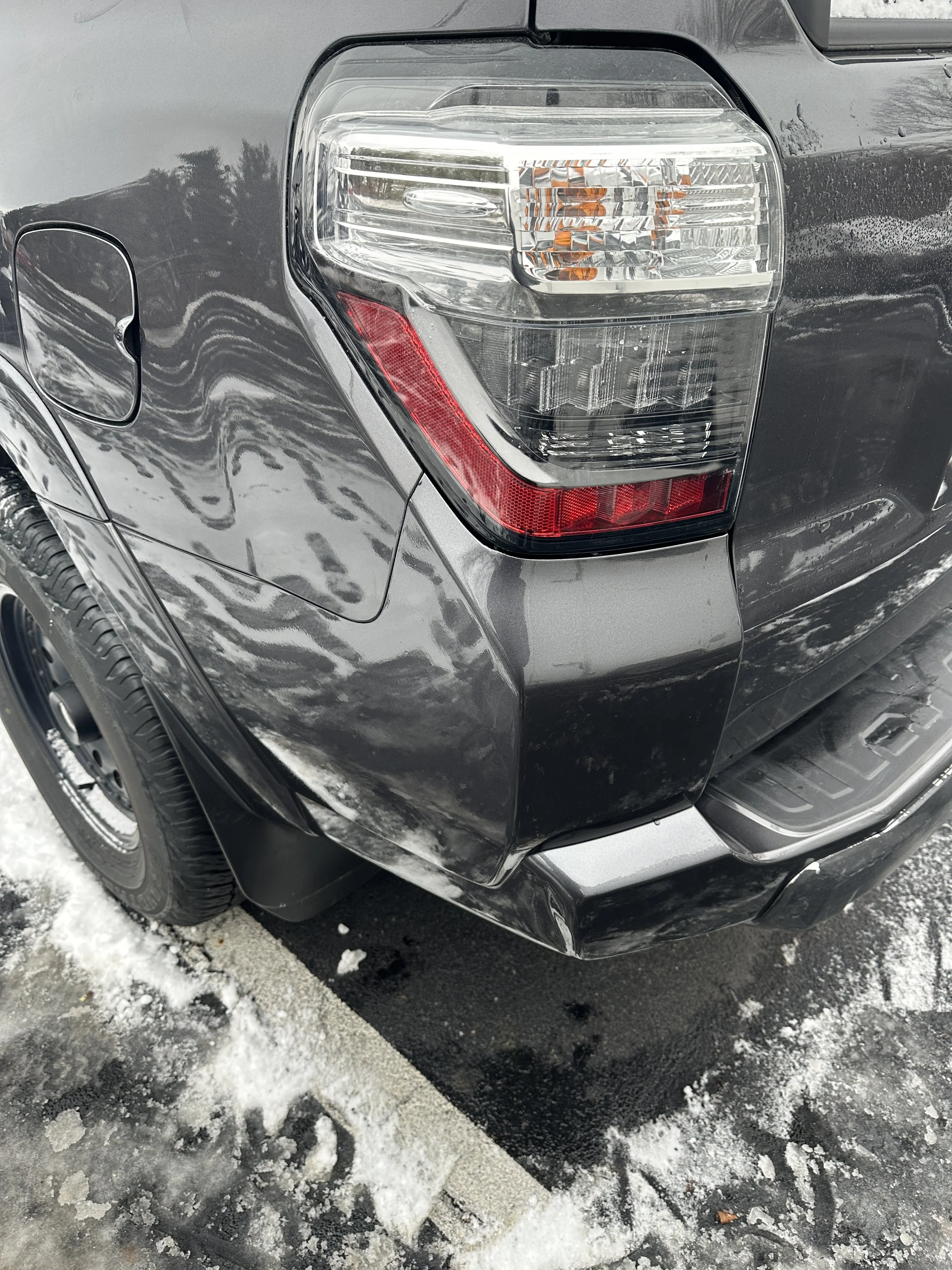Close-up of the rear corner of a black pickup truck parked on snow-covered pavement, showing the tail light, black wheel, and bumper.