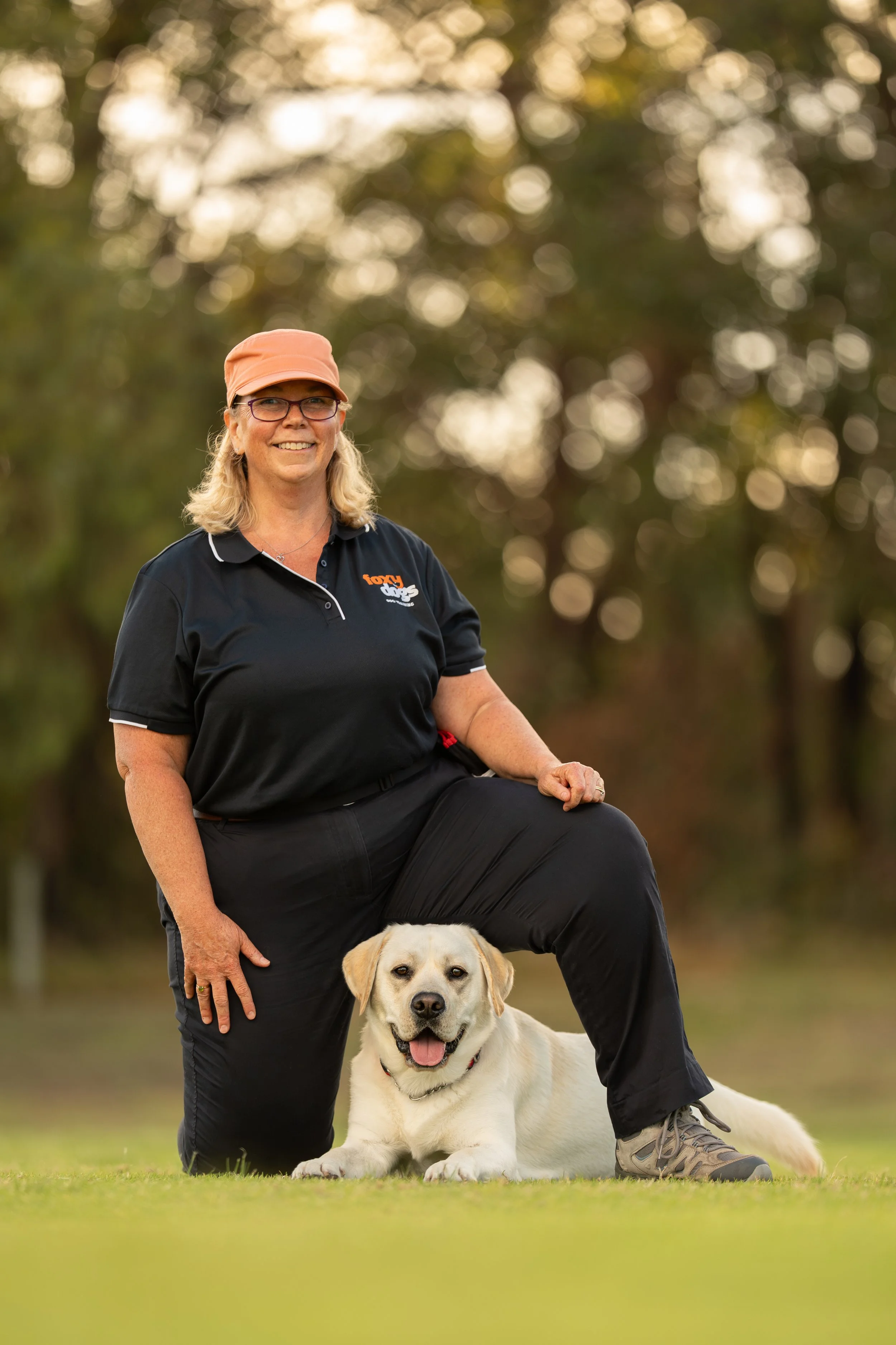 A woman with blonde hair, glasses, wearing a black polo shirt and pink cap, is kneeling outdoors with her Labrador Retriever in a grassy park area with trees in the background. The dog is lying on the grass, panting, and looking at the camera.