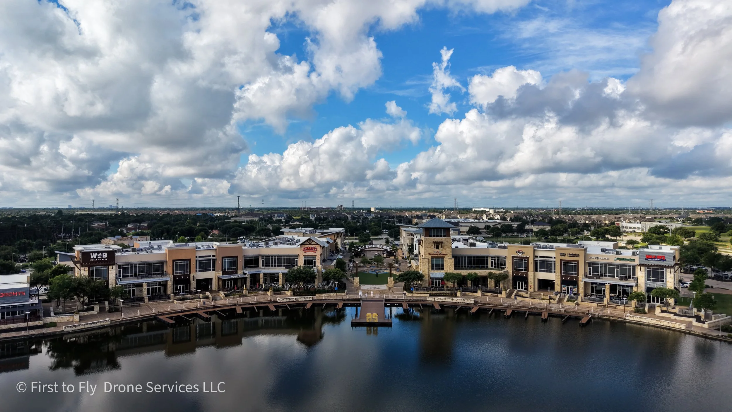 A view of a The Boardwalk in Towne Lake shopping center and a waterway with buildings and a cloudy sky in the background.