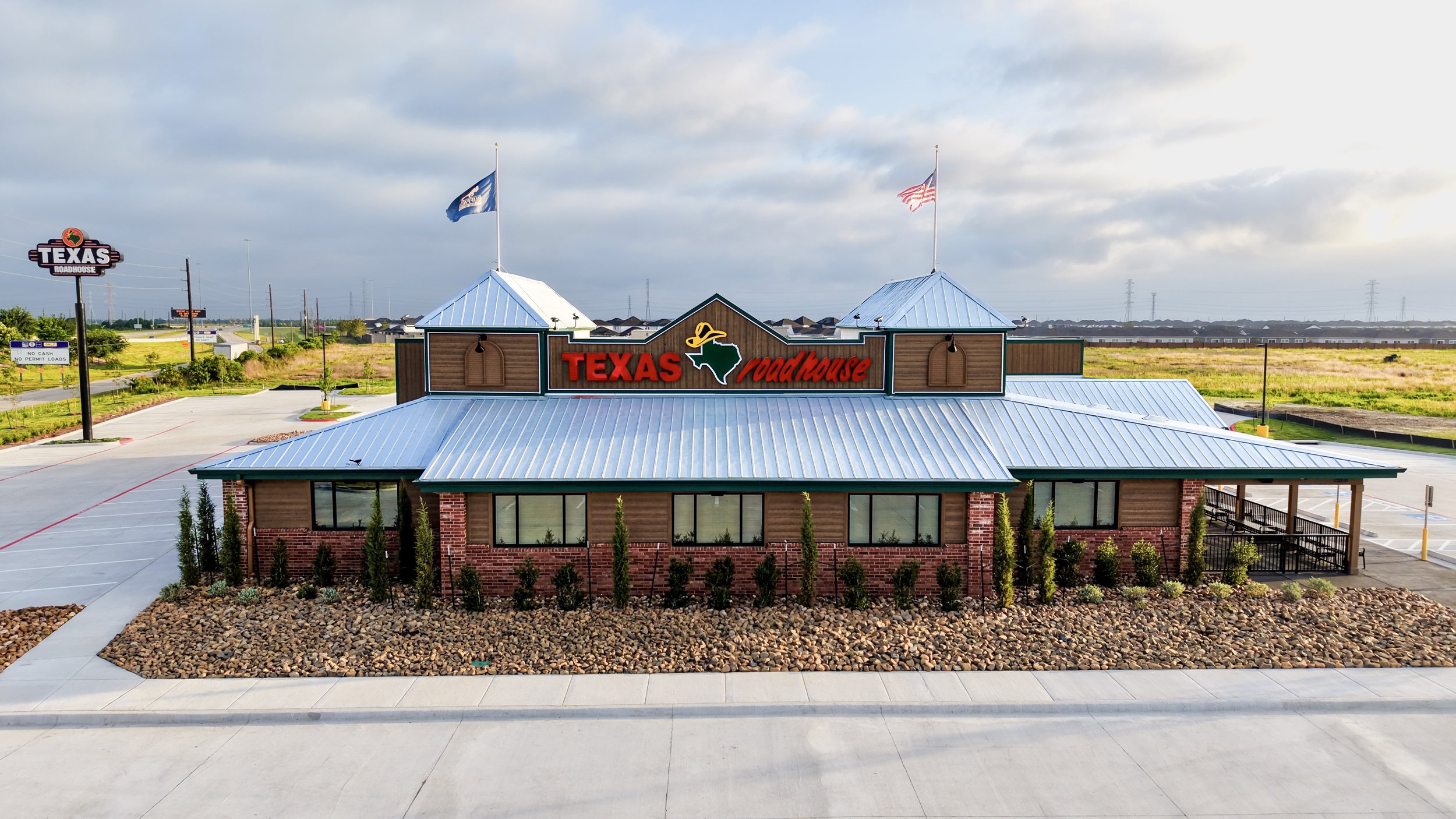 Exterior of a Texas Roadhouse restaurant with a brick and wood facade, blue metal roof, and large red sign with the restaurant's name and logo, surrounded by a parking lot and landscaping.